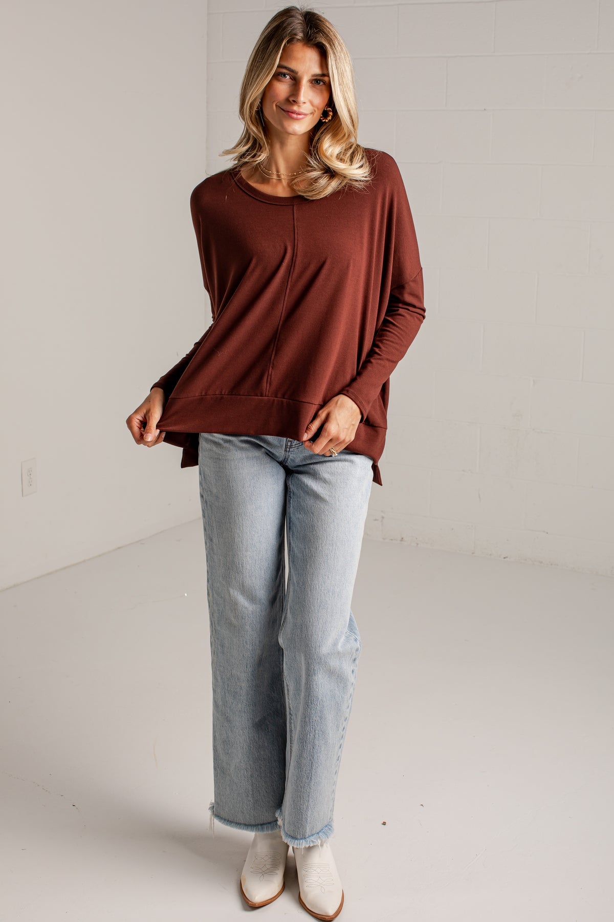 A woman modeling a rust-brown long-sleeve knit top with a crew neckline, paired with light-wash blue jeans and white ankle boots, standing against a white brick wall.