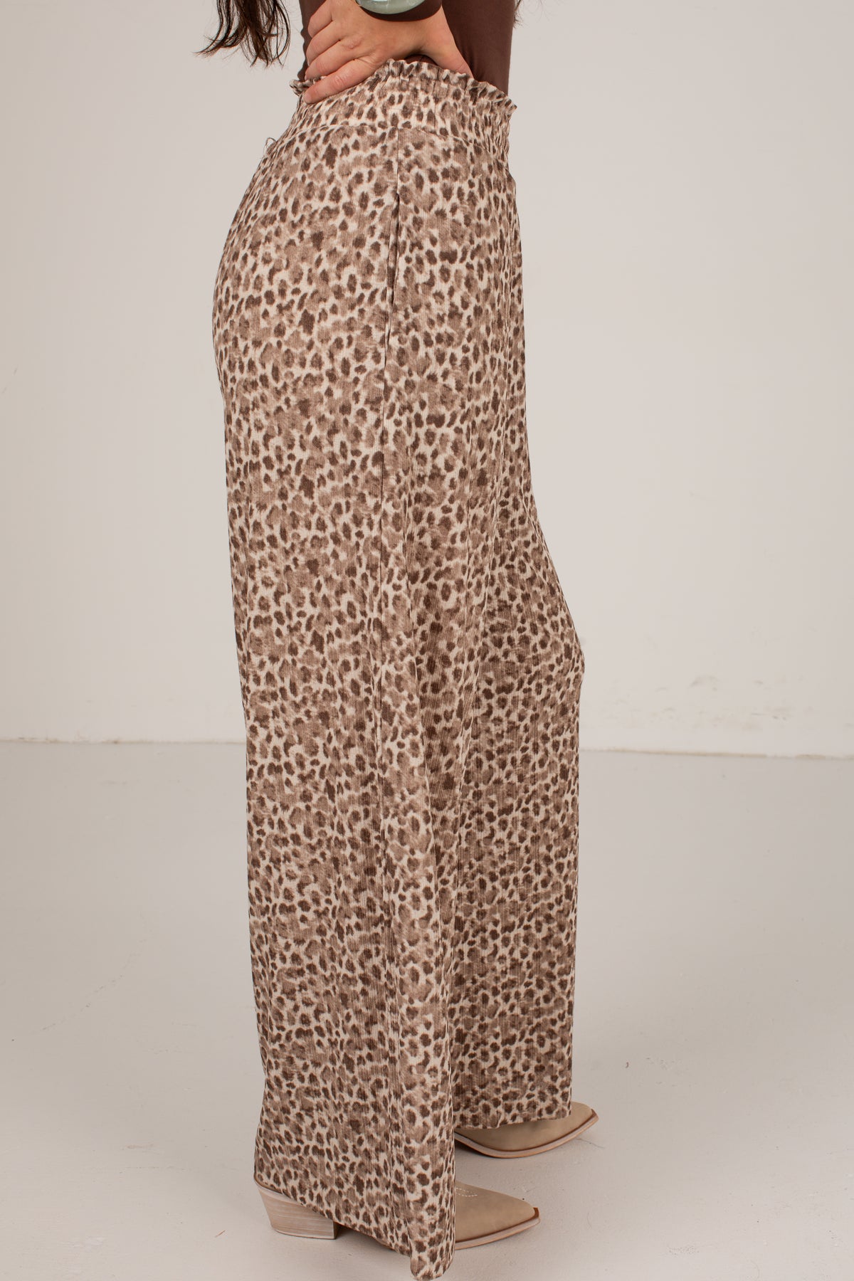 A person wearing high-waisted, wide-leg trousers with an allover leopard print pattern in beige, brown, and black, paired with beige block-heel mules against a plain off-white studio background.
