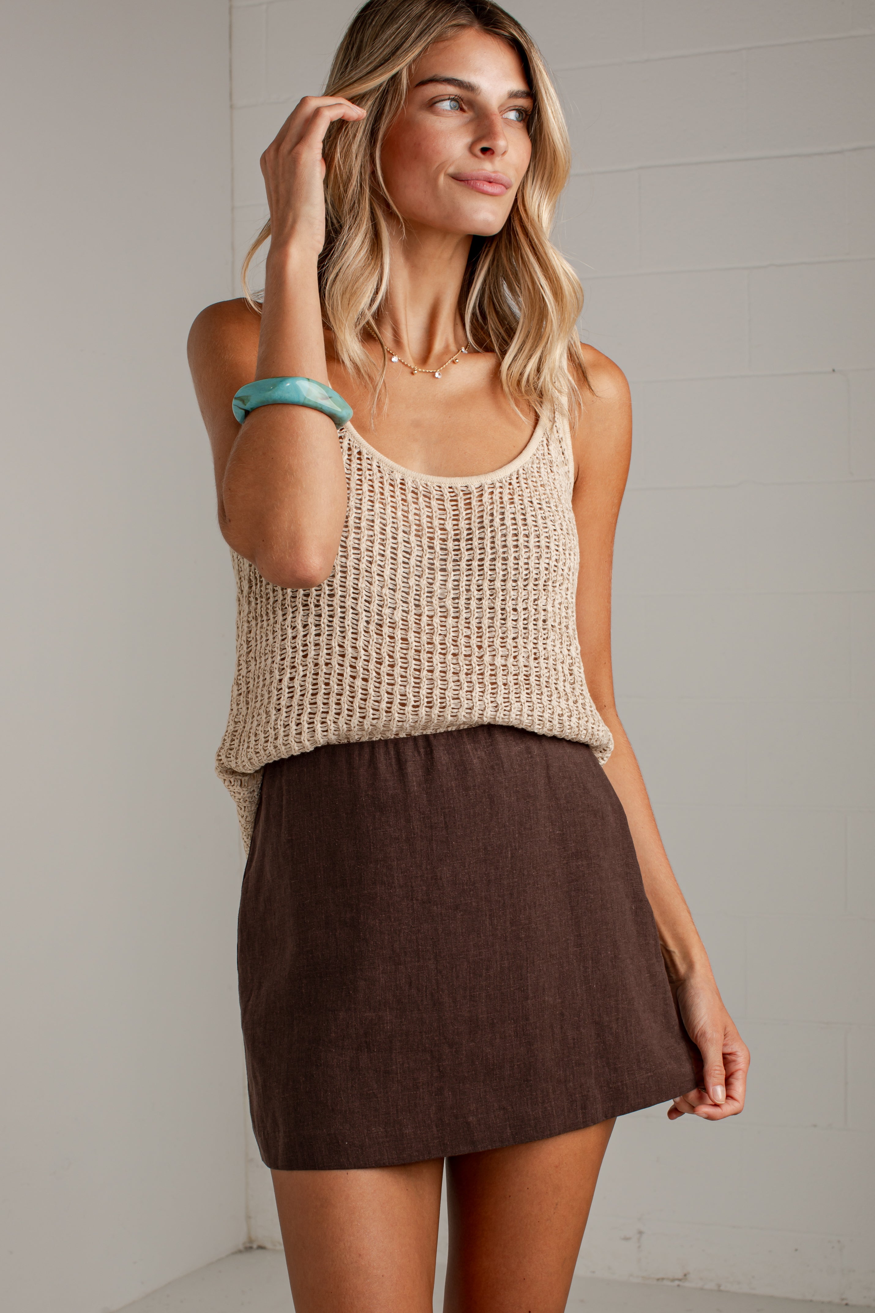 A woman modeling a beige chunky knit tank top and a brown linen mini skirt against a white paneled wall.
