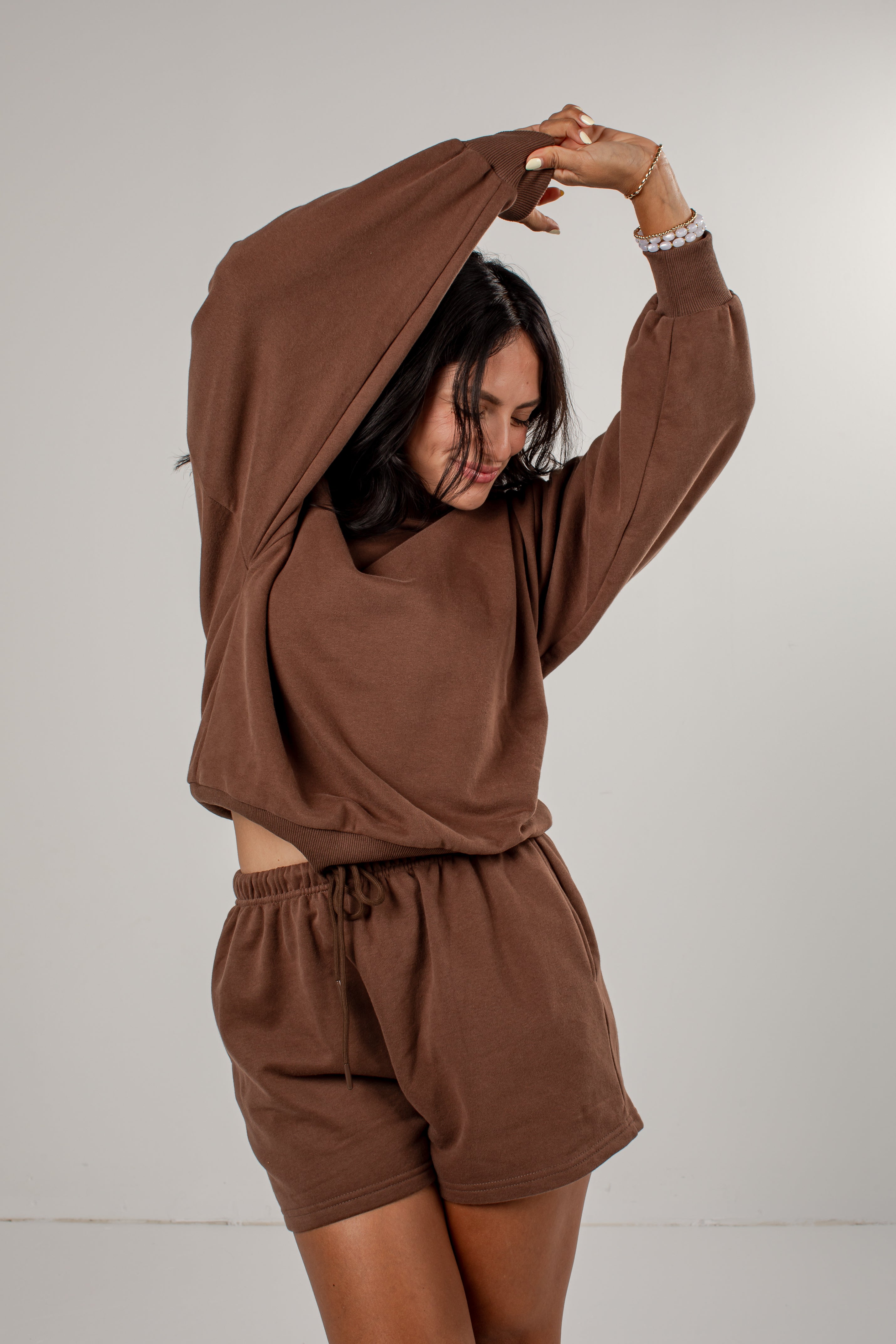 A woman modeling a matching brown lounge set consisting of a long-sleeve cropped sweatshirt and drawstring shorts, posed against a neutral light gray background.
