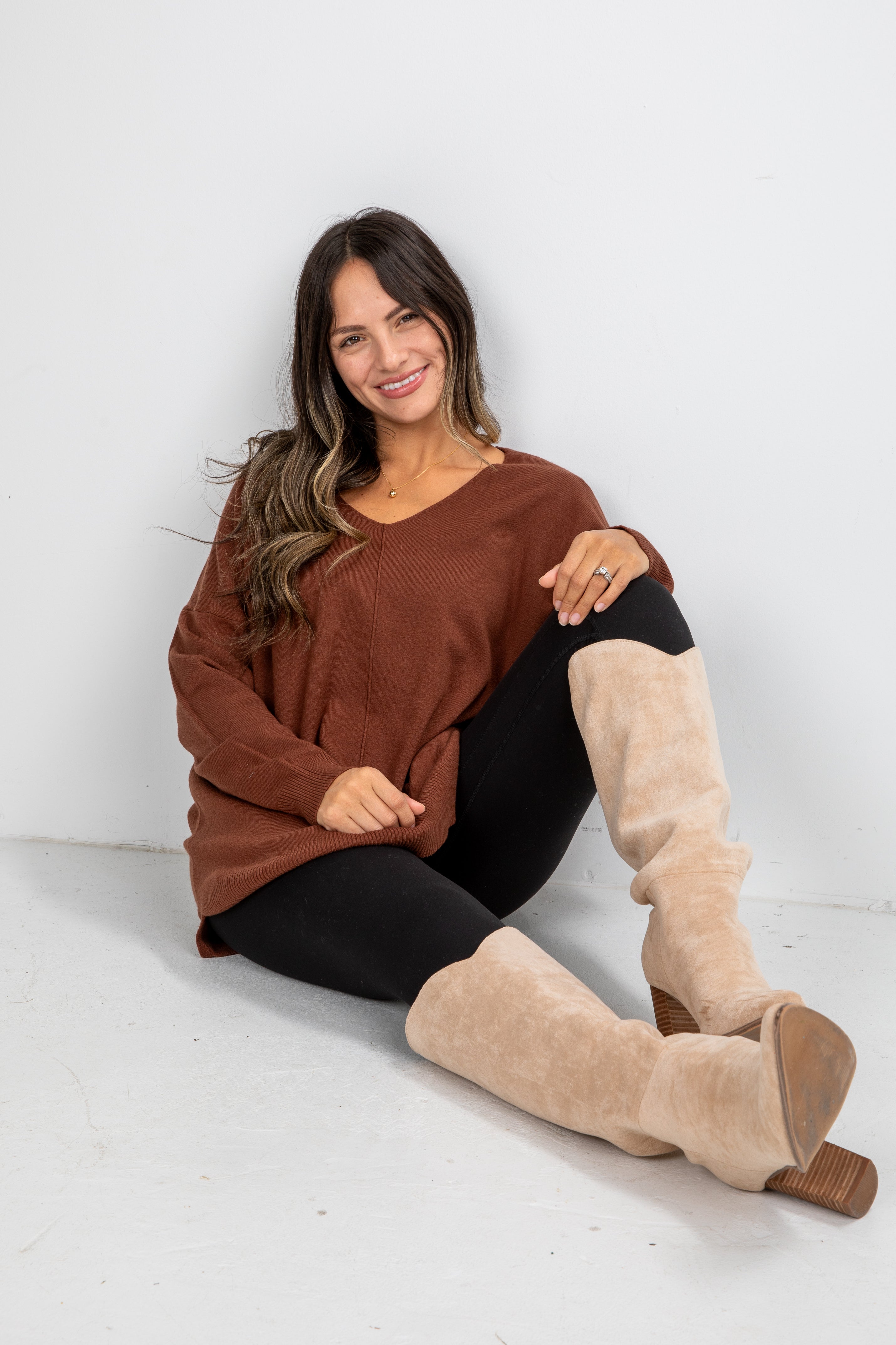 Woman wearing a brown sweater and beige knee-high boots sitting on a white floor.