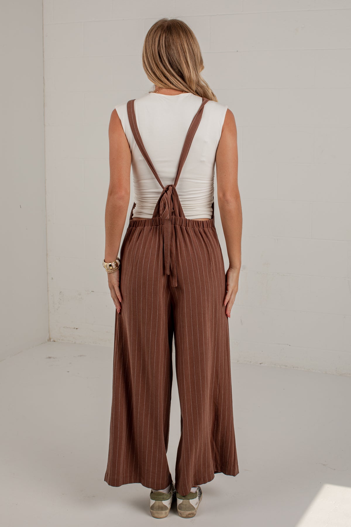 A woman viewed from behind wearing a sleeveless white top and brown wide-leg pleated pants with adjustable shoulder straps and a tie at the back, standing in a bright white studio.