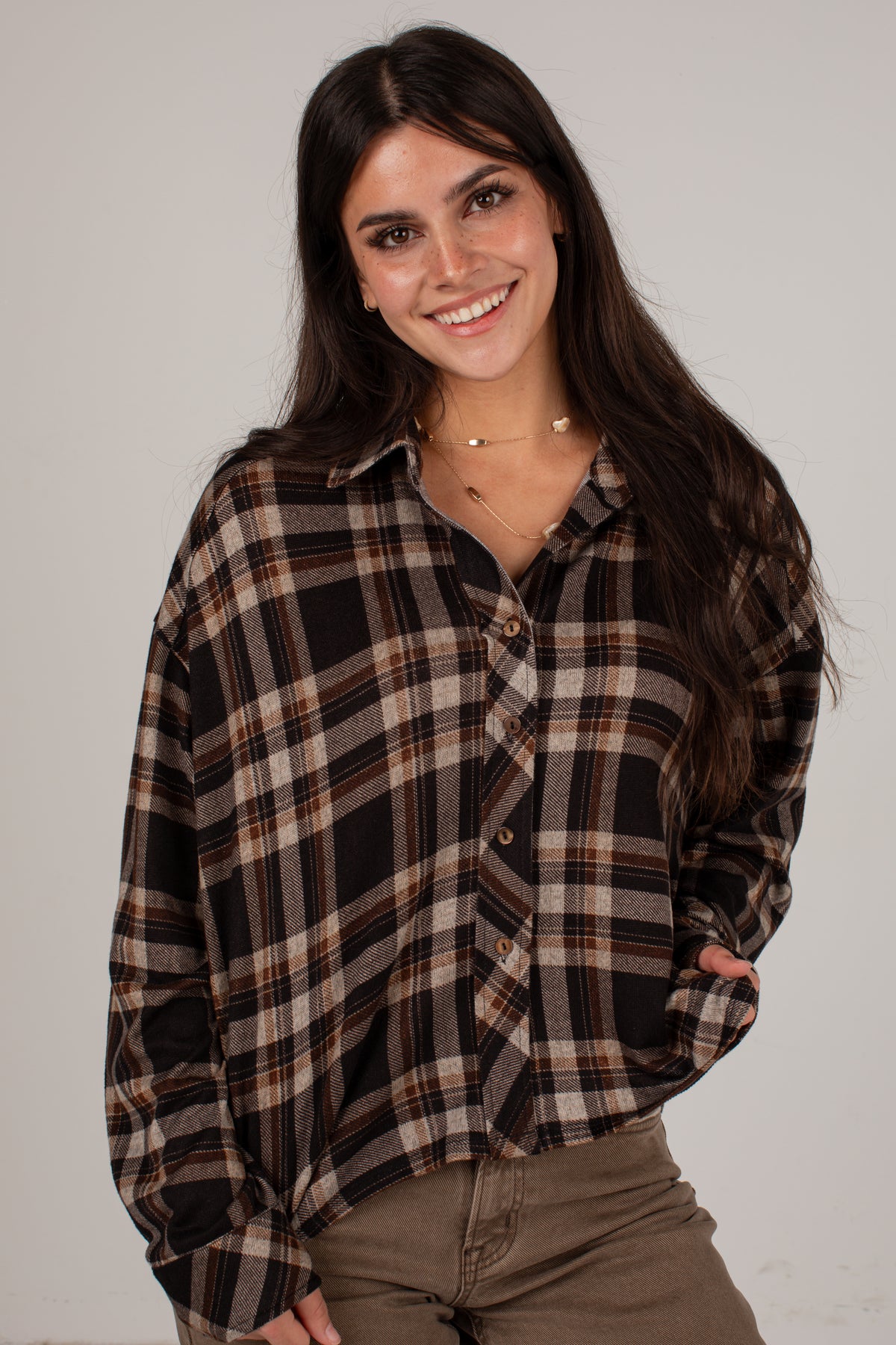 A woman with long dark hair smiling while wearing a brown, black, and beige plaid button-up shirt and brown pants, photographed against a plain light gray background.
