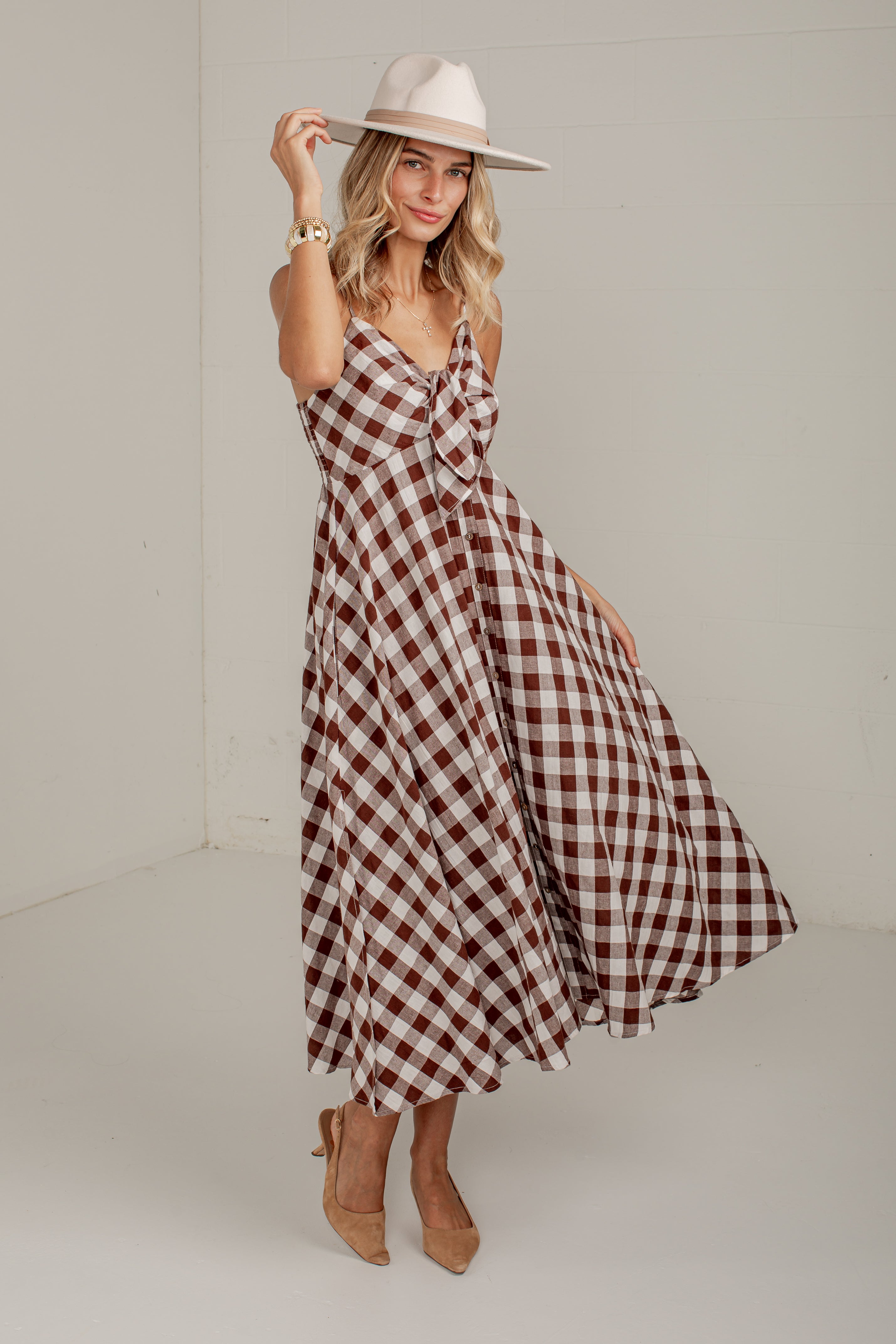 A woman modeling a brown and white gingham midi dress with a knotted bodice, styled with a wide-brimmed cream hat and tan heels in a neutral studio setting.