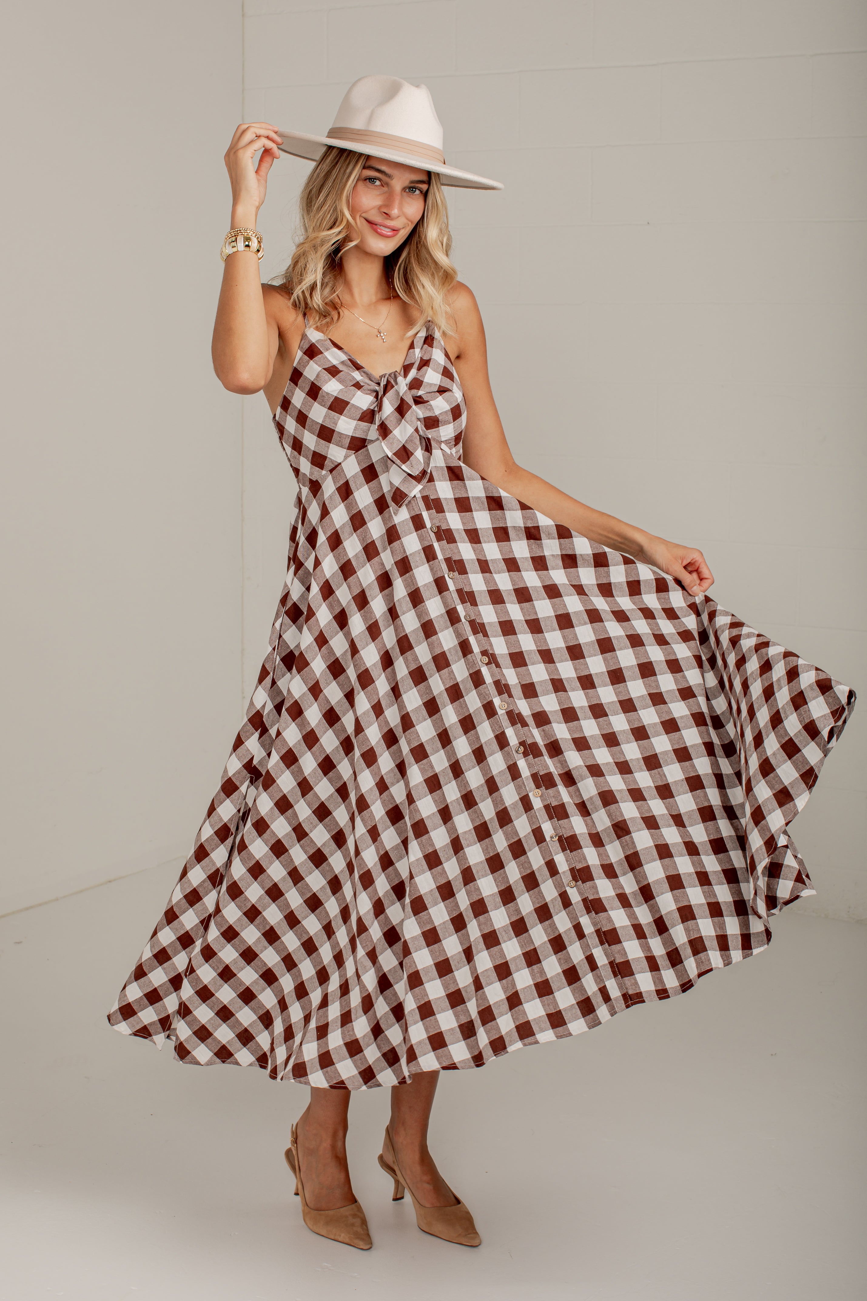 A woman modeling a brown and white gingham midi dress with spaghetti straps and a sweetheart neckline, paired with a cream wide-brim hat and tan heels, set against a plain light gray studio background.