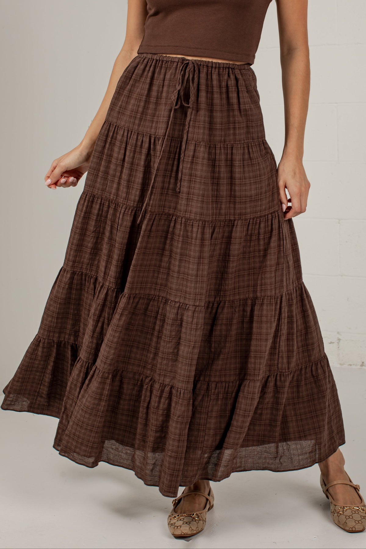A woman wearing a brown tiered maxi skirt with a subtle plaid pattern, paired with a matching brown short-sleeve crop top and brown flat shoes, standing against a light gray wall.