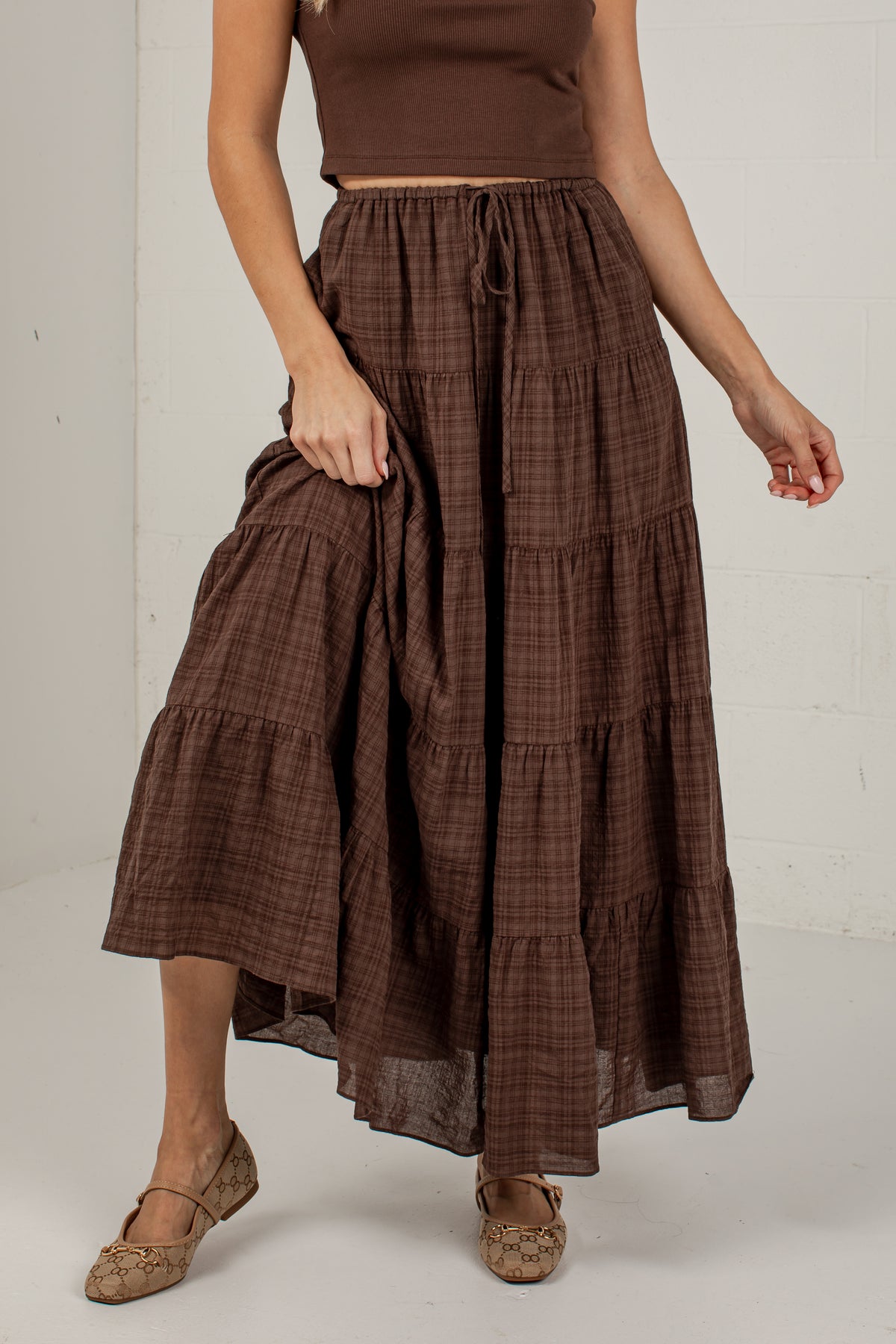 A model wearing a brown tiered maxi skirt with a subtle plaid pattern, paired with a matching brown crop top and brown flat shoes, standing against a white wall.