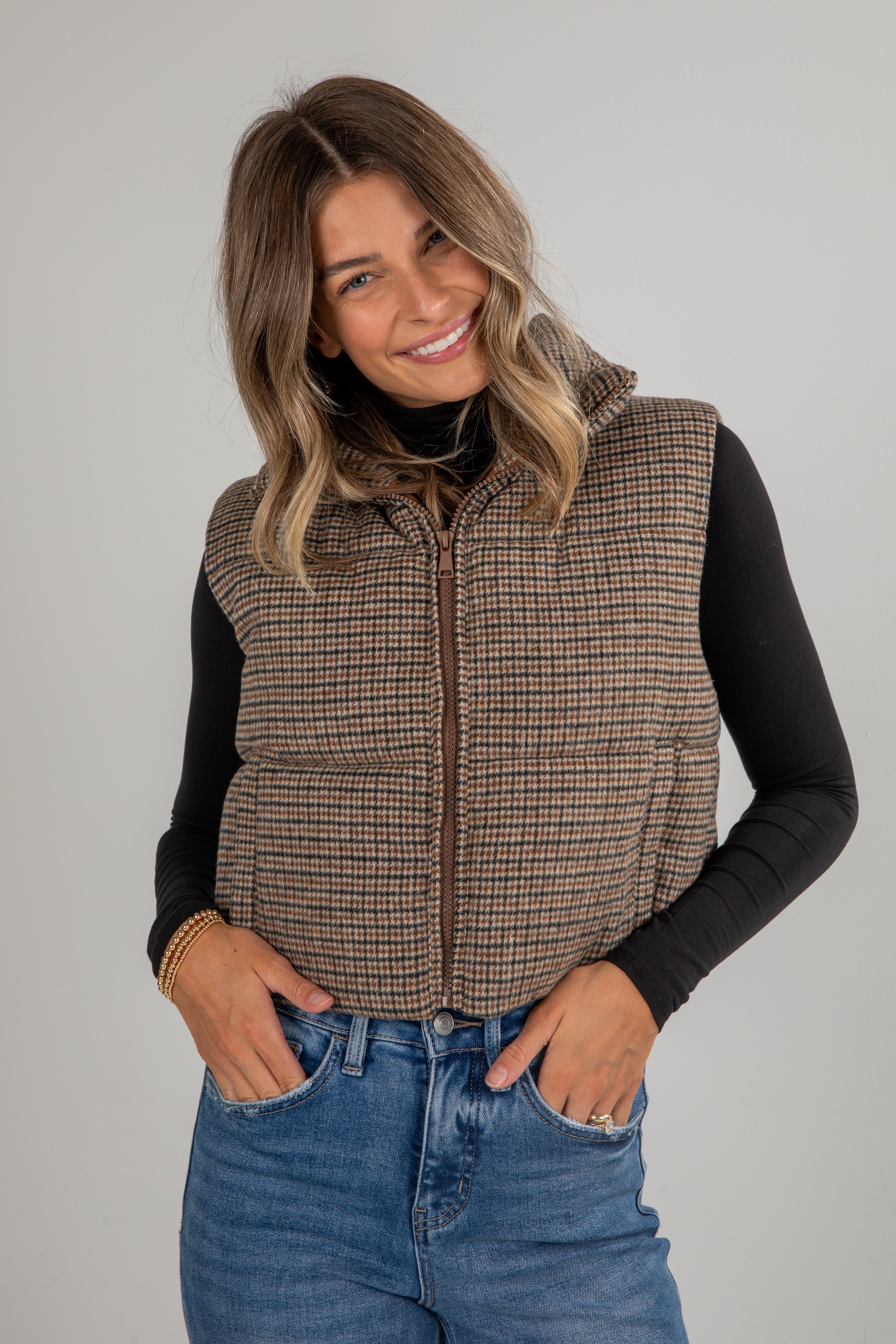 Woman wearing a plaid vest over a black long-sleeve shirt and blue jeans on a gray background