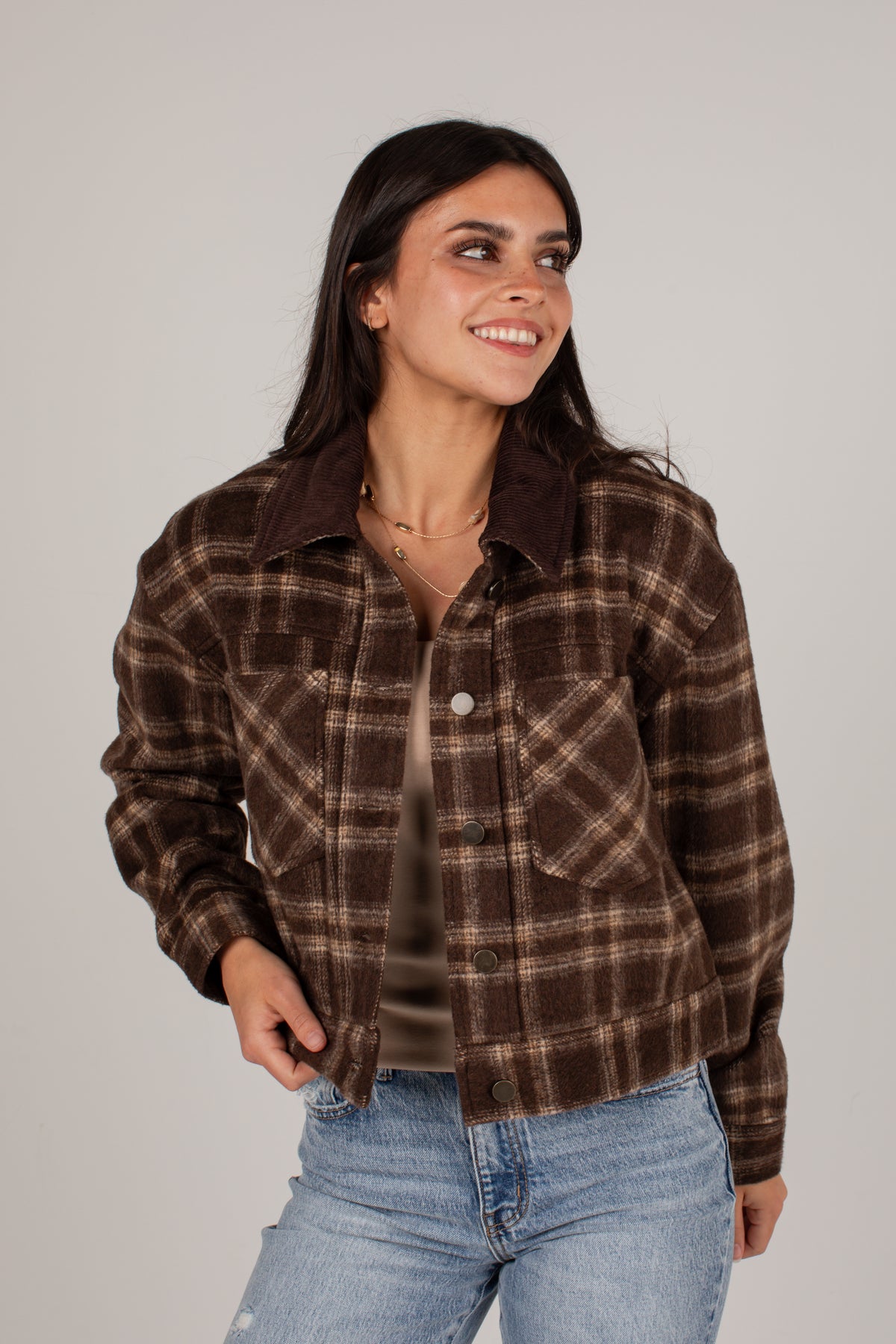 A woman wearing a brown and beige plaid flannel button-up jacket with a corduroy collar, paired with a brown top and light blue jeans, photographed against a solid light gray background.