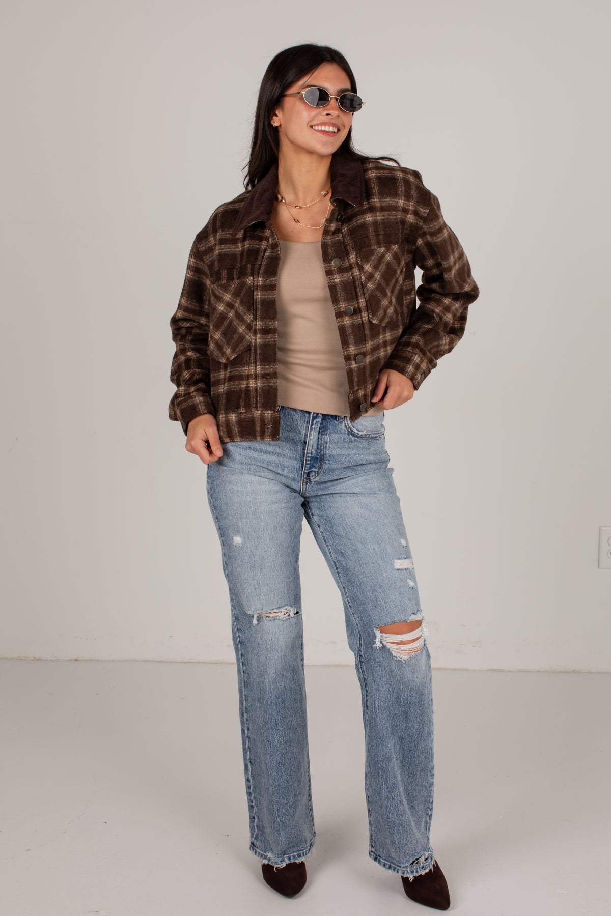 A woman modeling a brown and black plaid flannel shirt jacket over a beige tank top and light-wash distressed jeans, standing against a plain off-white studio background.
