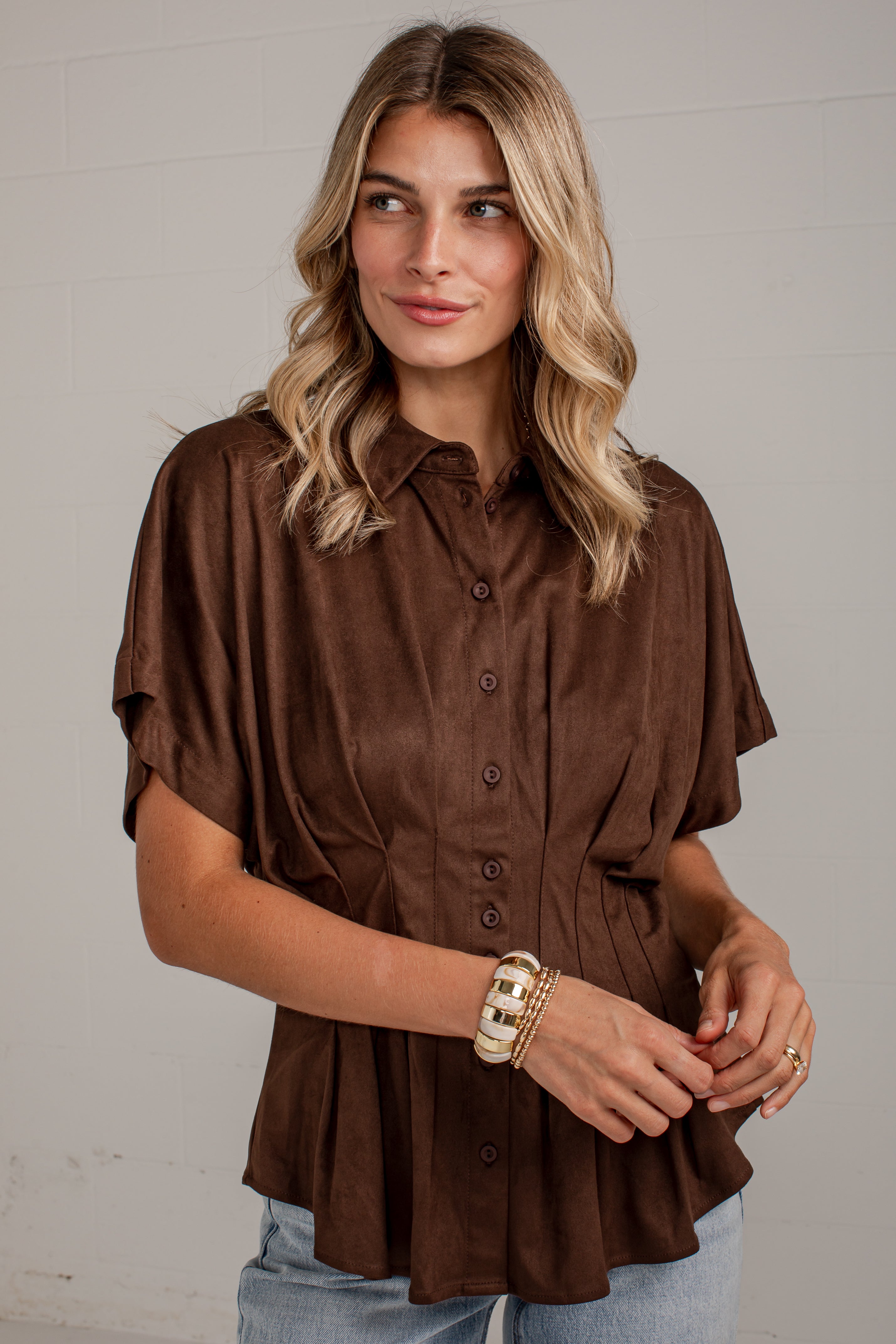 A woman wearing a brown short-sleeve button-up blouse with a gathered peplum hem, paired with light blue jeans, standing against a light gray brick wall.