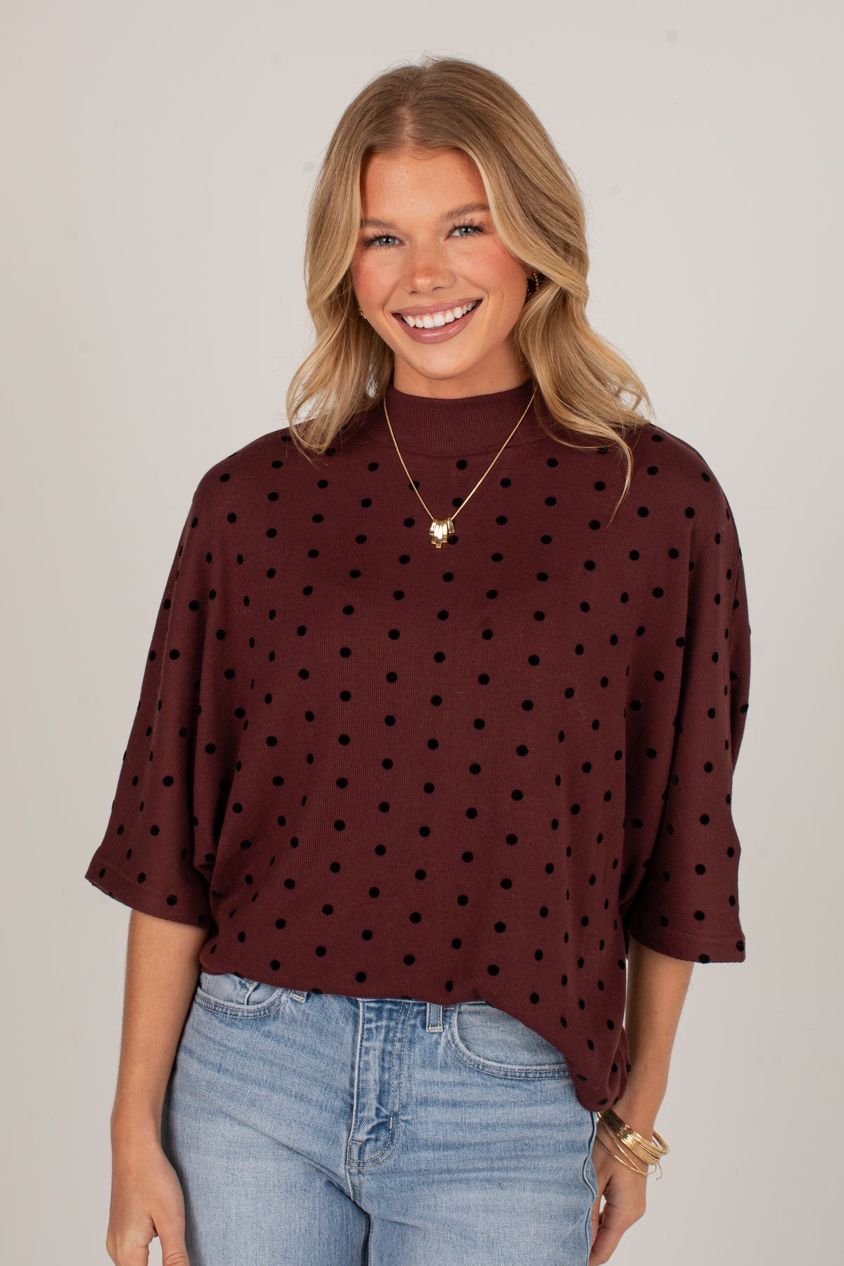 A smiling blonde woman wearing a dark brown mock neck top with black polka dots and light blue jeans, standing against a plain light gray studio background.