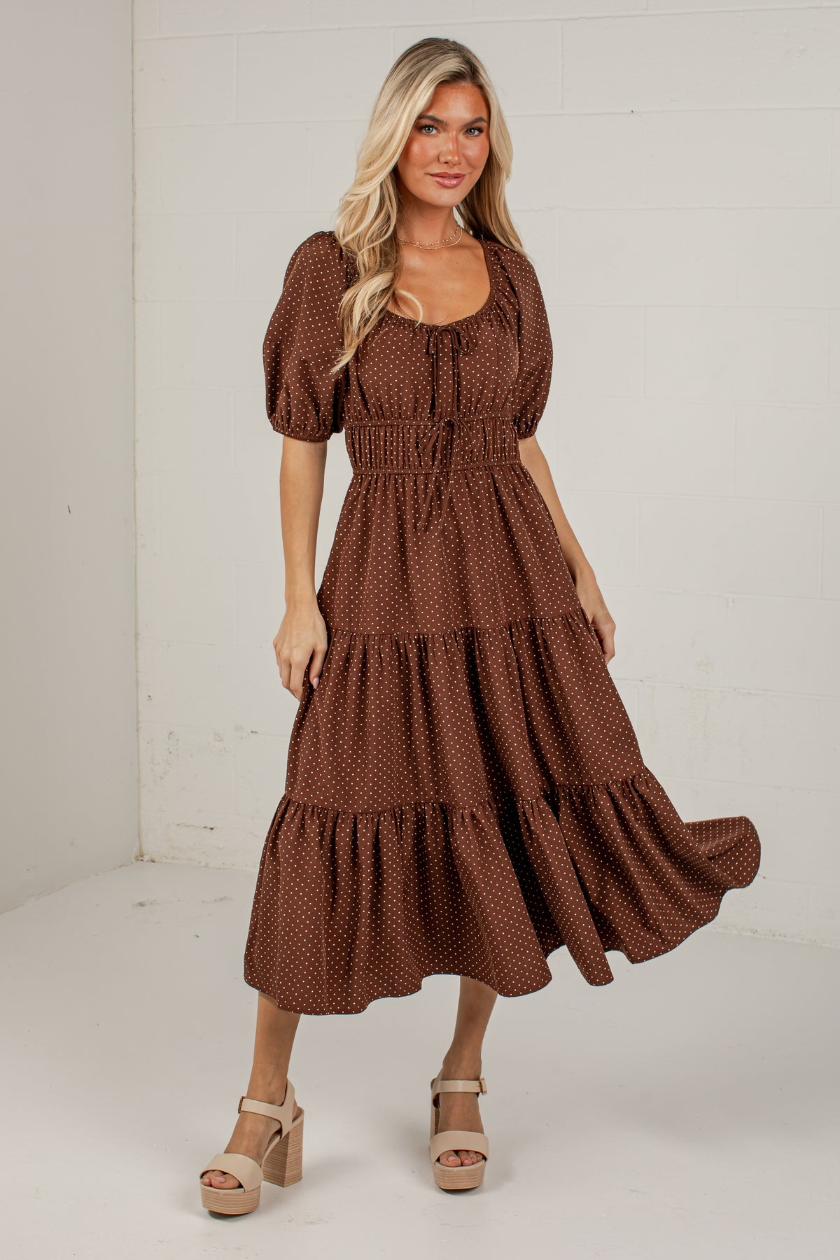 A woman modeling a brown tiered midi dress with a small white polka dot pattern, puff sleeves, and a smocked waist, standing against a white brick wall.