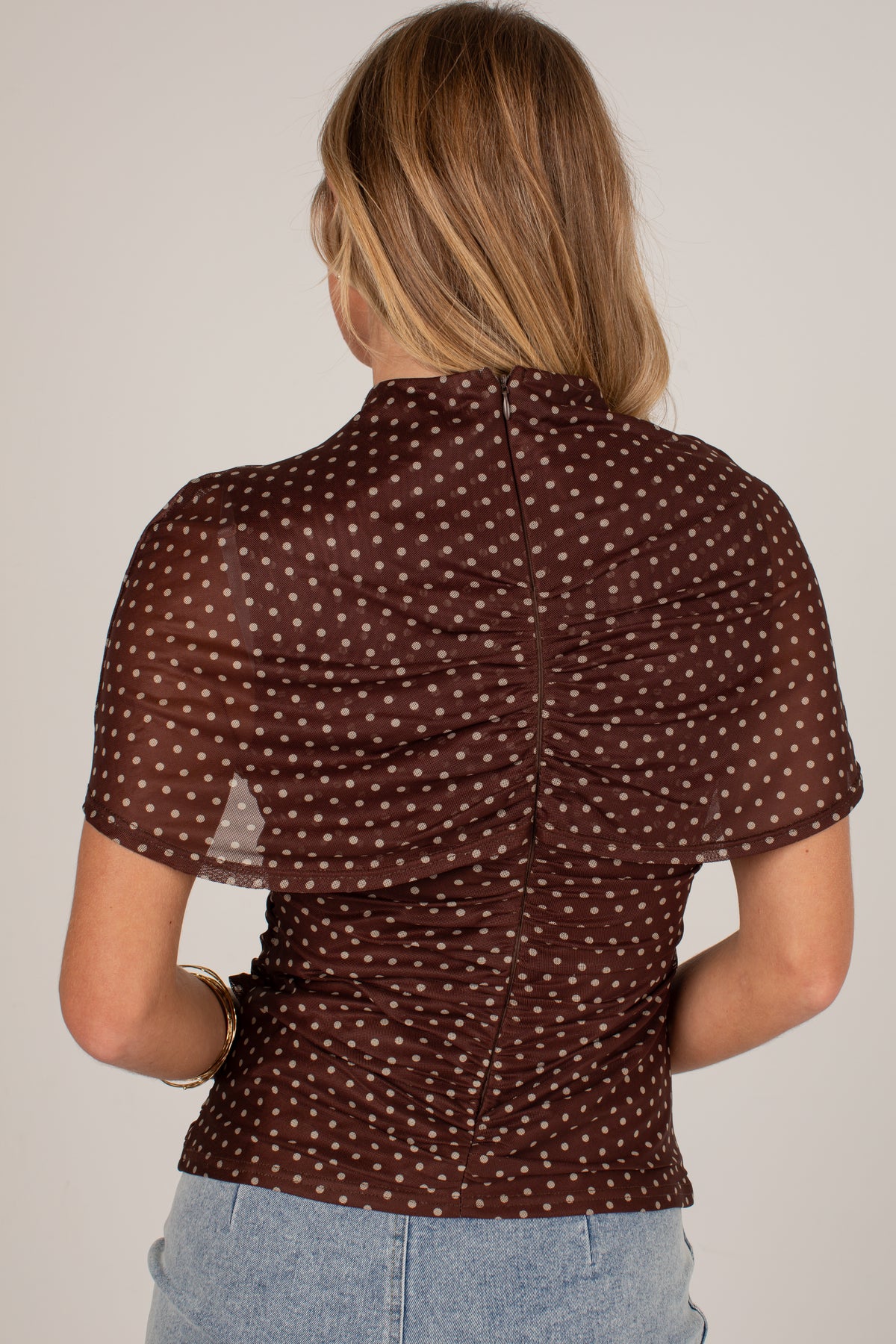 A woman viewed from the back wearing a brown sheer short-sleeve top with a white polka dot pattern and gathered ruching at the back, paired with light blue jeans against a plain light gray background.