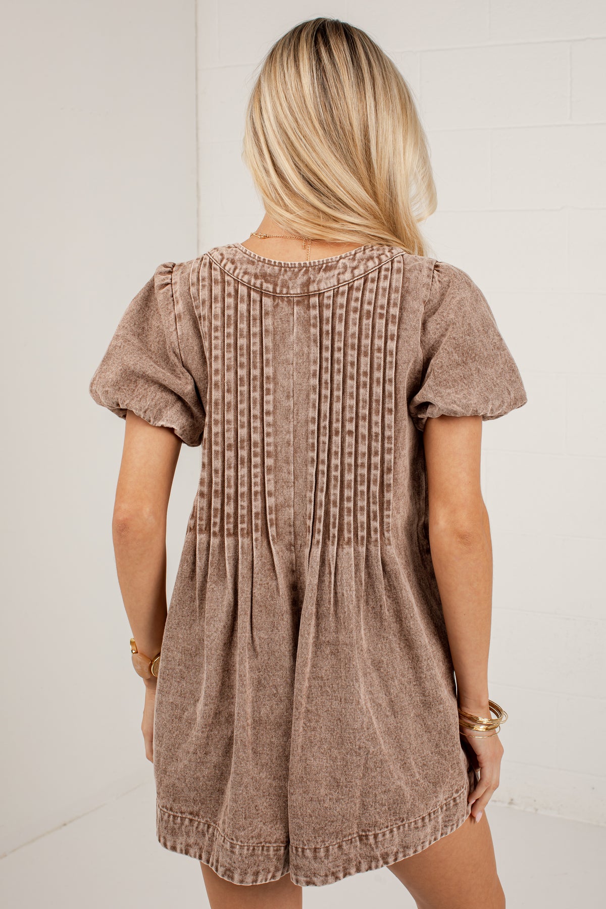 A woman viewed from behind wearing a short-sleeve brown romper with vertical textured stitching on the back, set against a plain white wall.