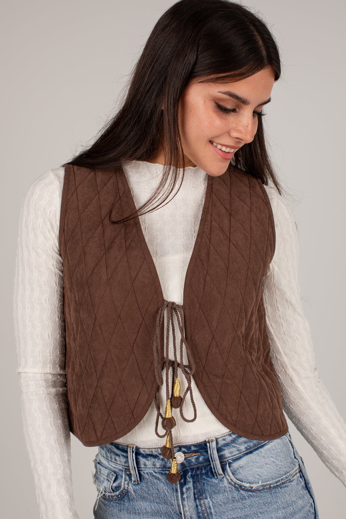 A woman wearing a brown diamond-quilted vest over a white long-sleeve top and blue jeans, photographed against a plain light gray studio background.