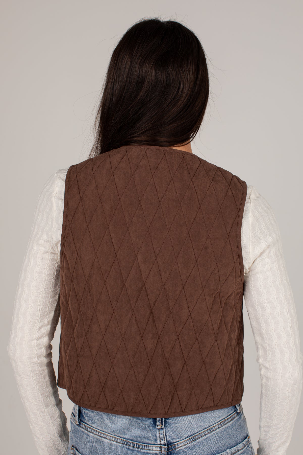 A person shown from the back wearing a brown diamond-quilted sleeveless vest over a white ribbed long-sleeve top and light blue jeans, against a plain light gray studio background.