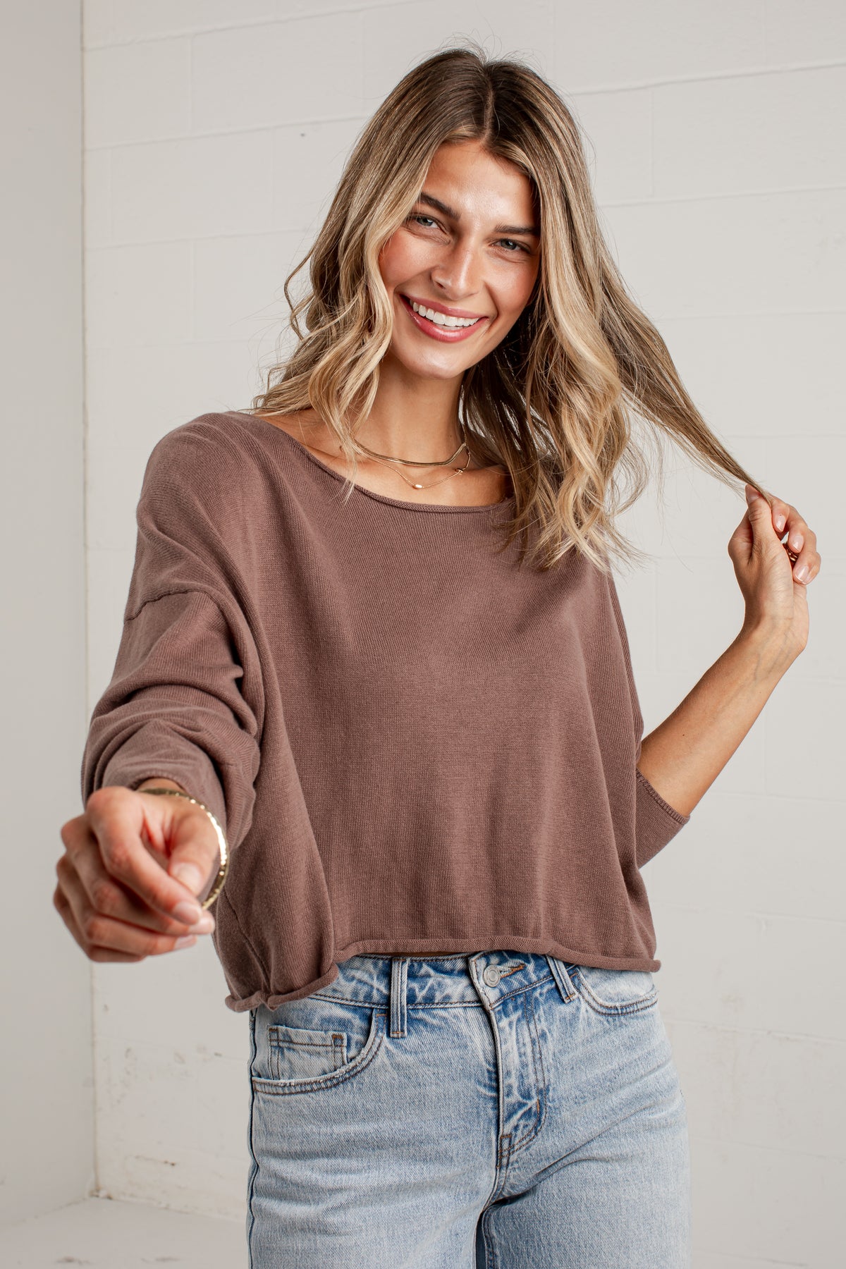 A woman wearing a brown long-sleeve cropped knit top and light blue jeans, smiling and pointing toward the camera against a white paneled wall.