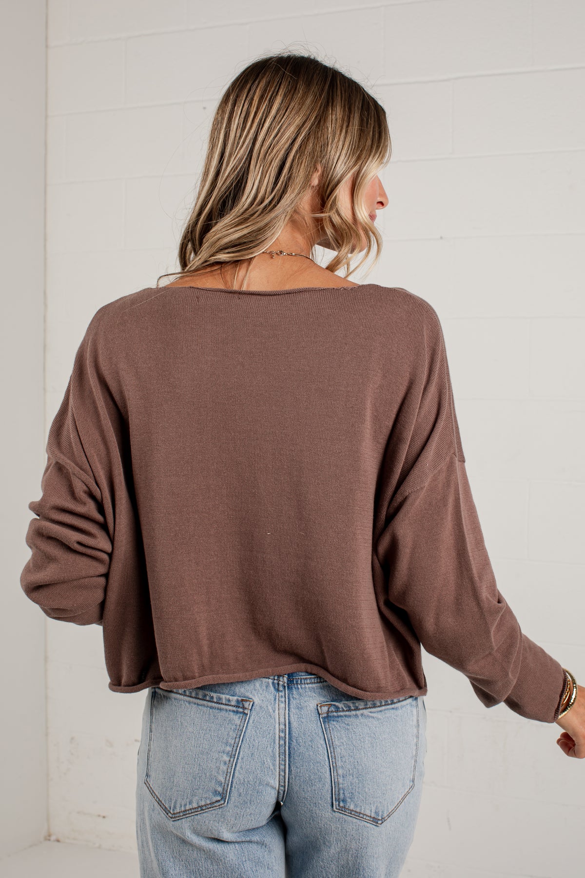 A woman viewed from behind wearing a brown long-sleeve knit top and light blue jeans, standing against a white paneled wall.
