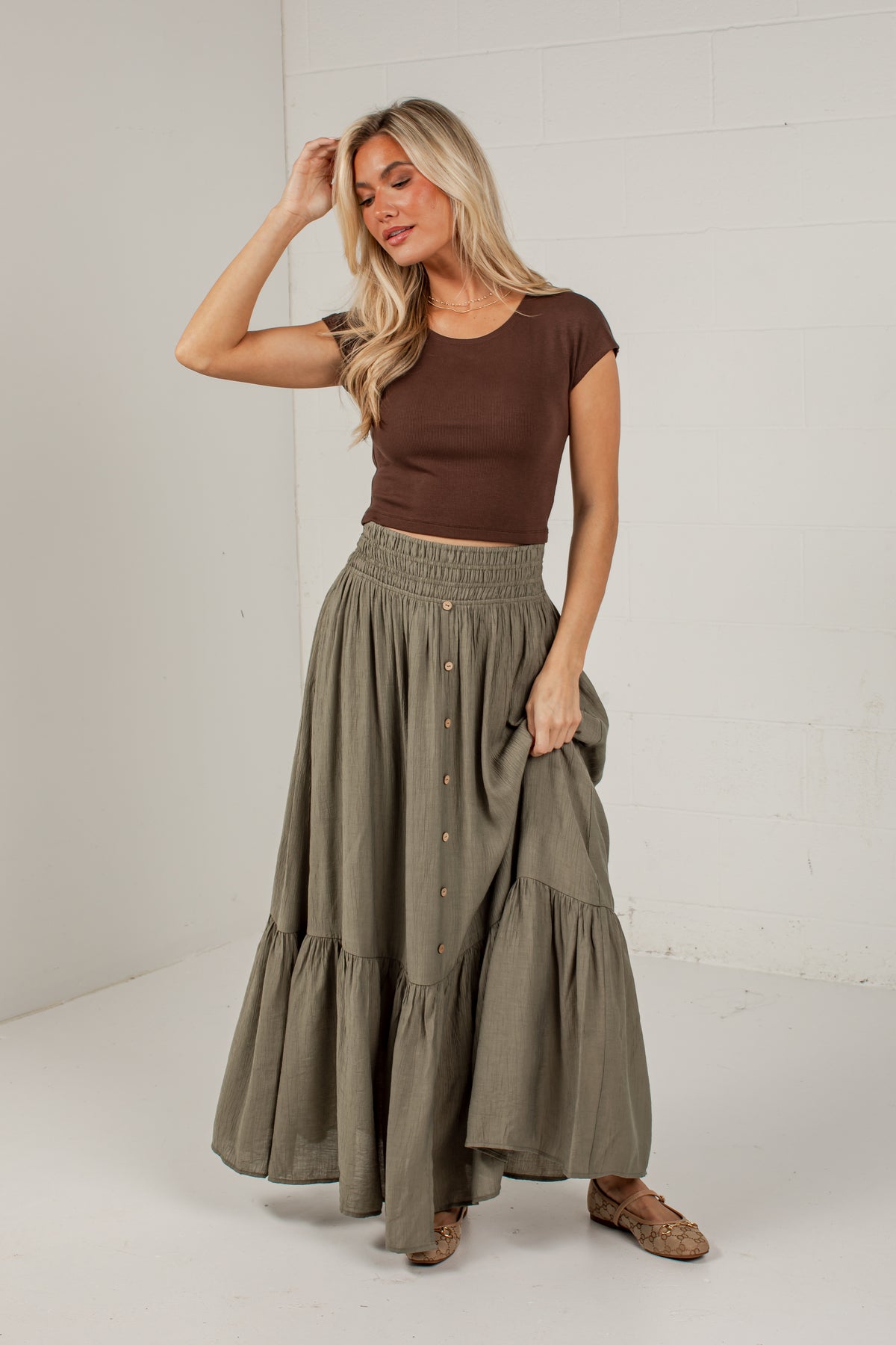 A woman modeling a brown short-sleeve crop top and an olive green tiered maxi skirt with front buttons, standing against a white brick wall.
