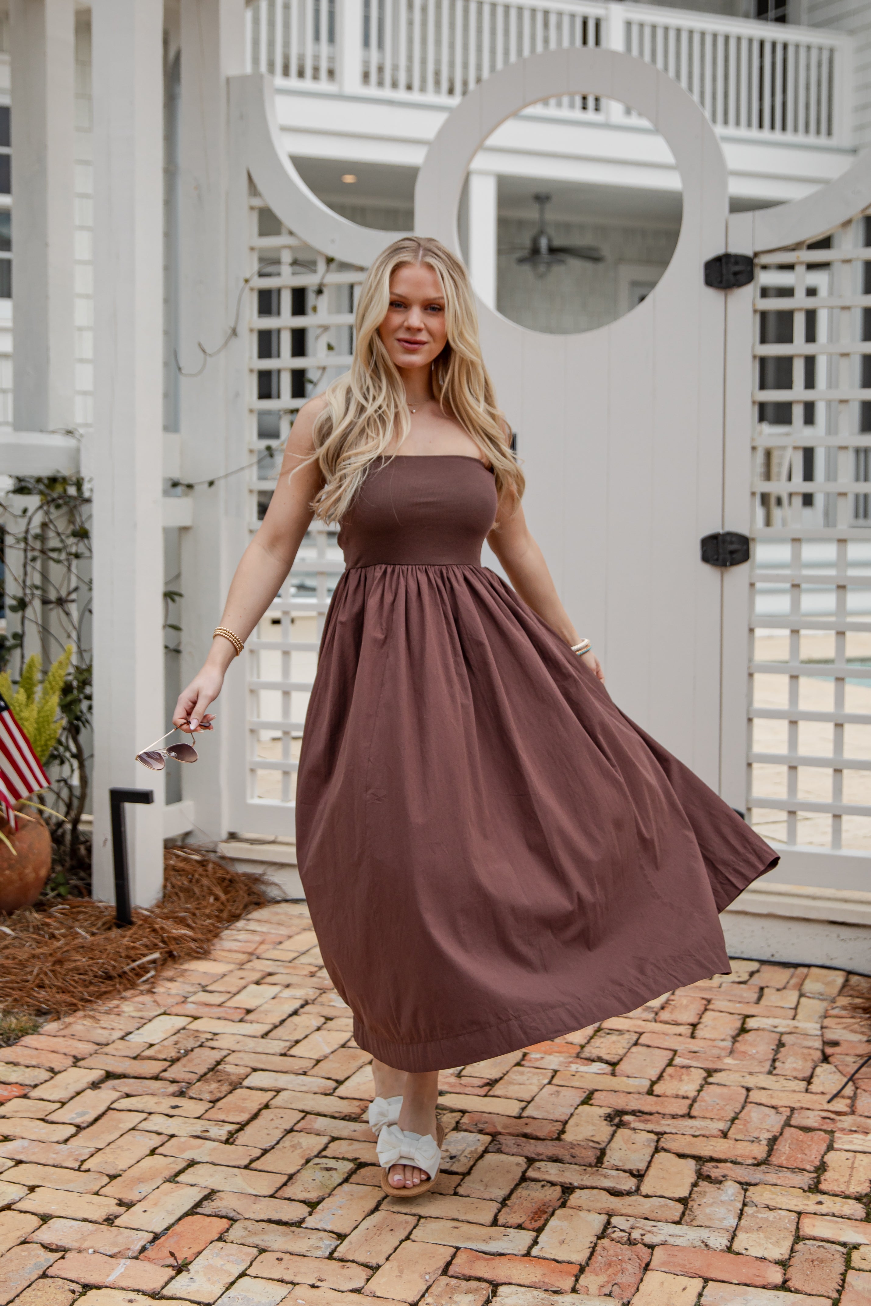 Woman in a brown dress standing on a brick patio with a white house in the background