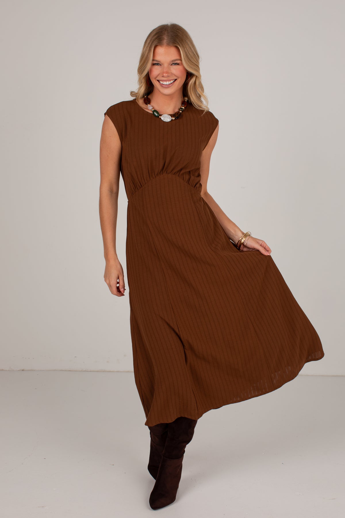 A woman modeling a sleeveless, ribbed brown midi dress with a gathered waist, paired with dark brown boots, against a plain light gray studio background.