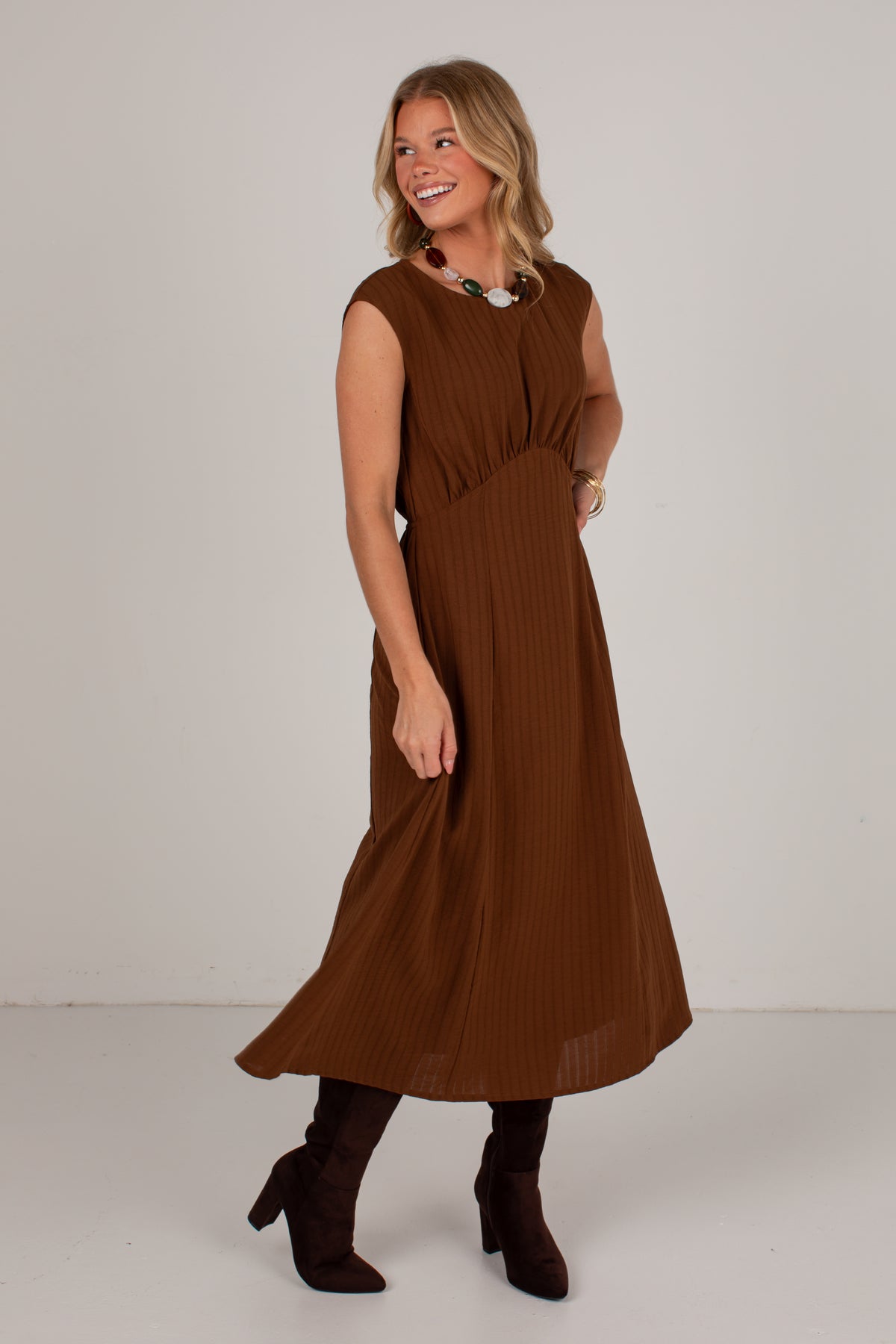 A woman modeling a sleeveless brown midi dress with a gathered waist and pleated texture, paired with dark brown ankle boots, against a plain light gray studio background.