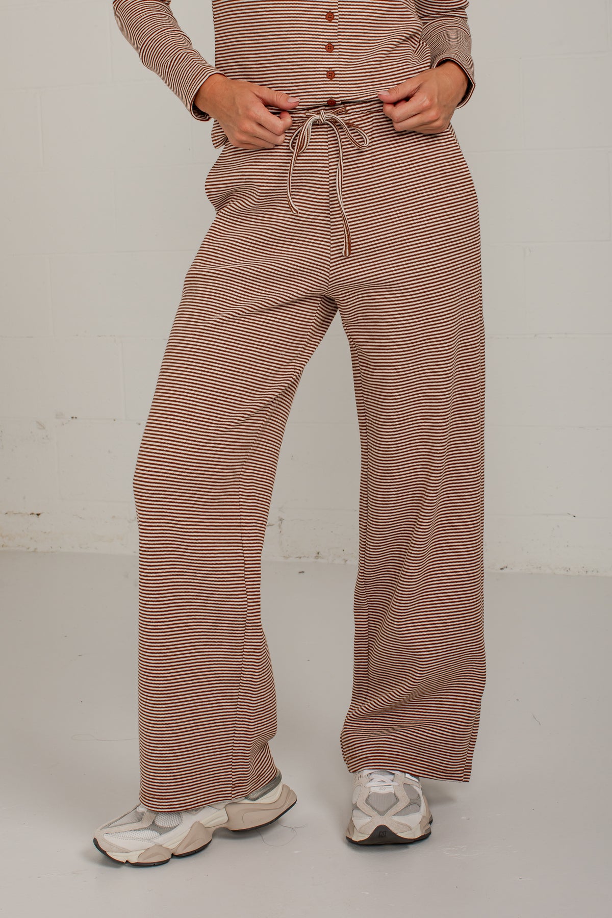 A person wearing loose, wide-leg pants and a matching long-sleeve top with thin brown and beige horizontal stripes, standing against a plain light wall.