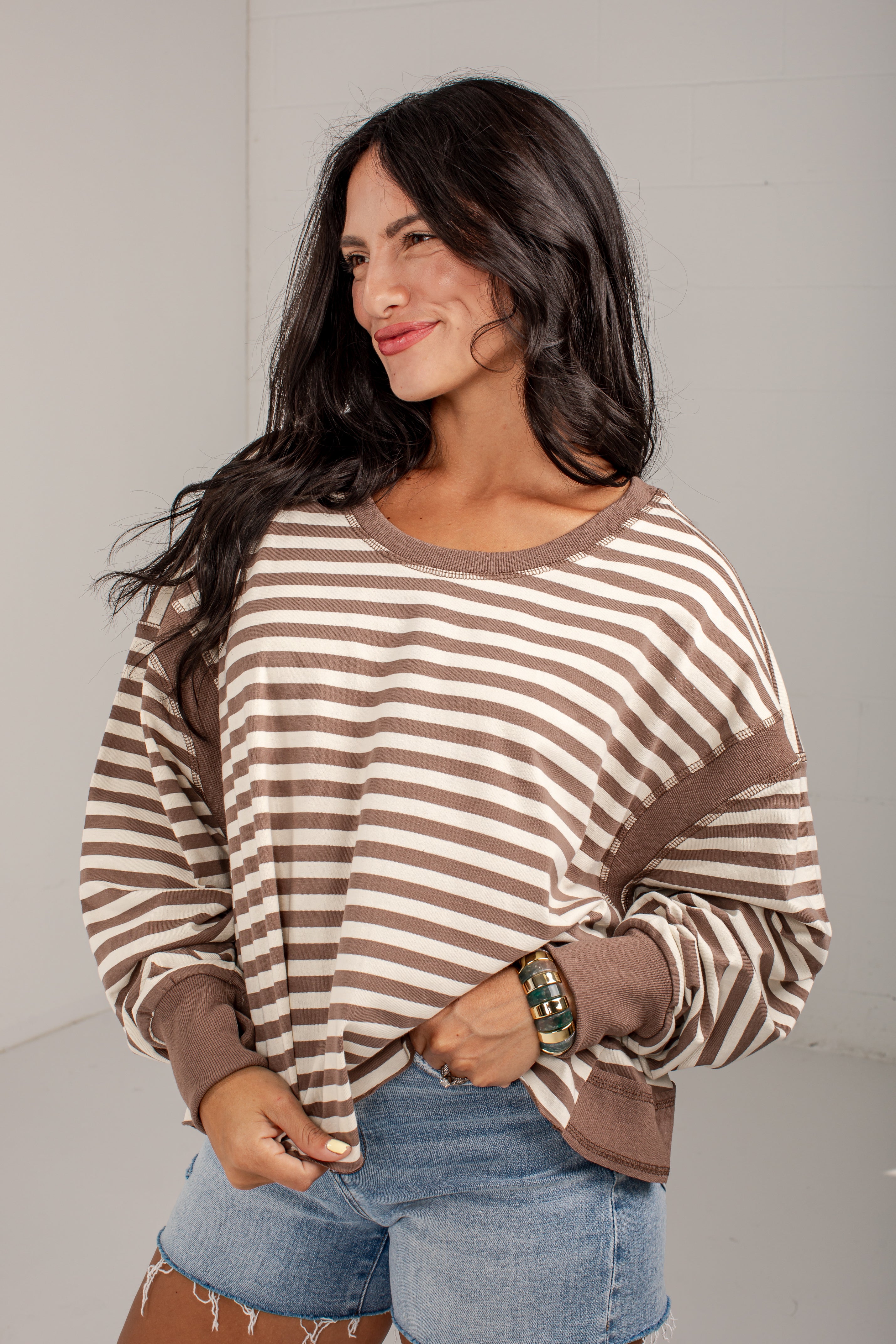 A woman wearing a brown and beige horizontally striped long-sleeve top with solid brown ribbed trim, paired with denim shorts, standing against a plain light gray wall.
