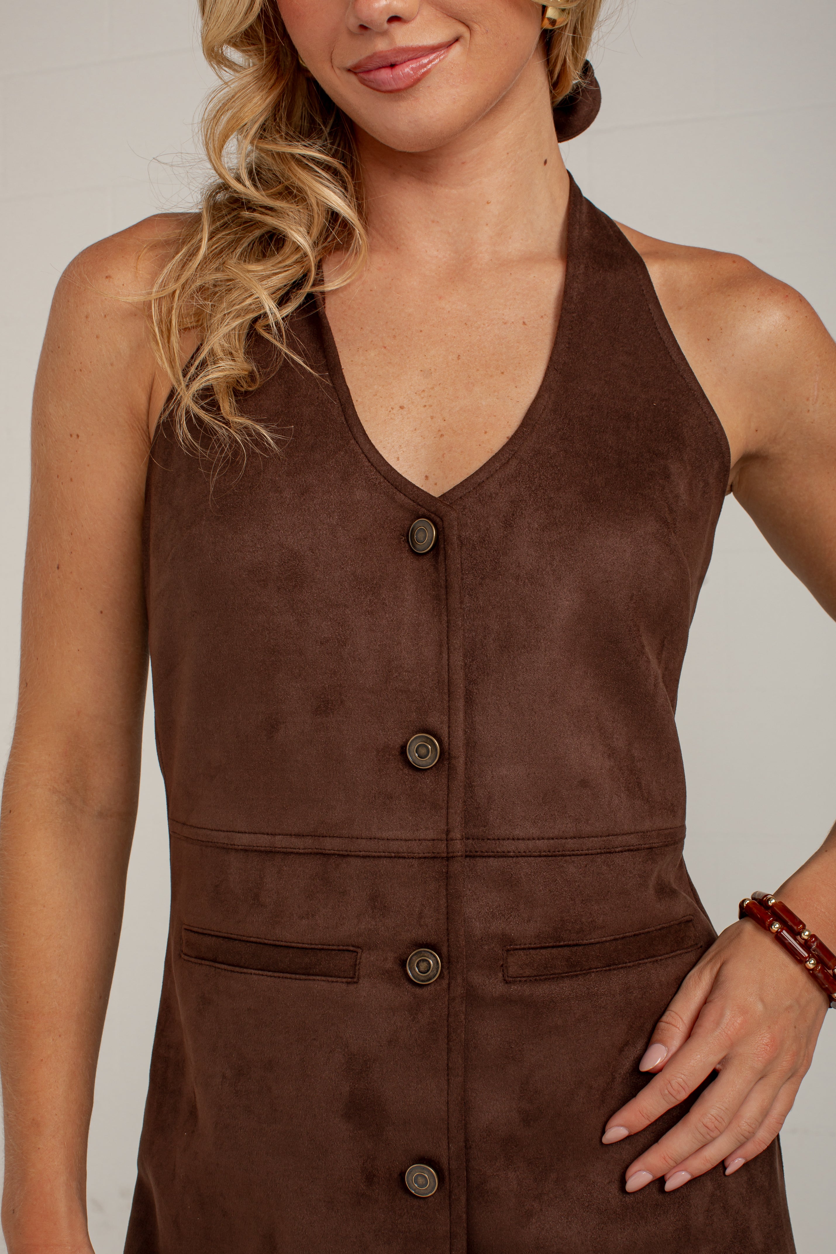 A close-up of a woman wearing a sleeveless brown faux-suede halter top with a button-front closure and front patch pockets, photographed against a plain light background.