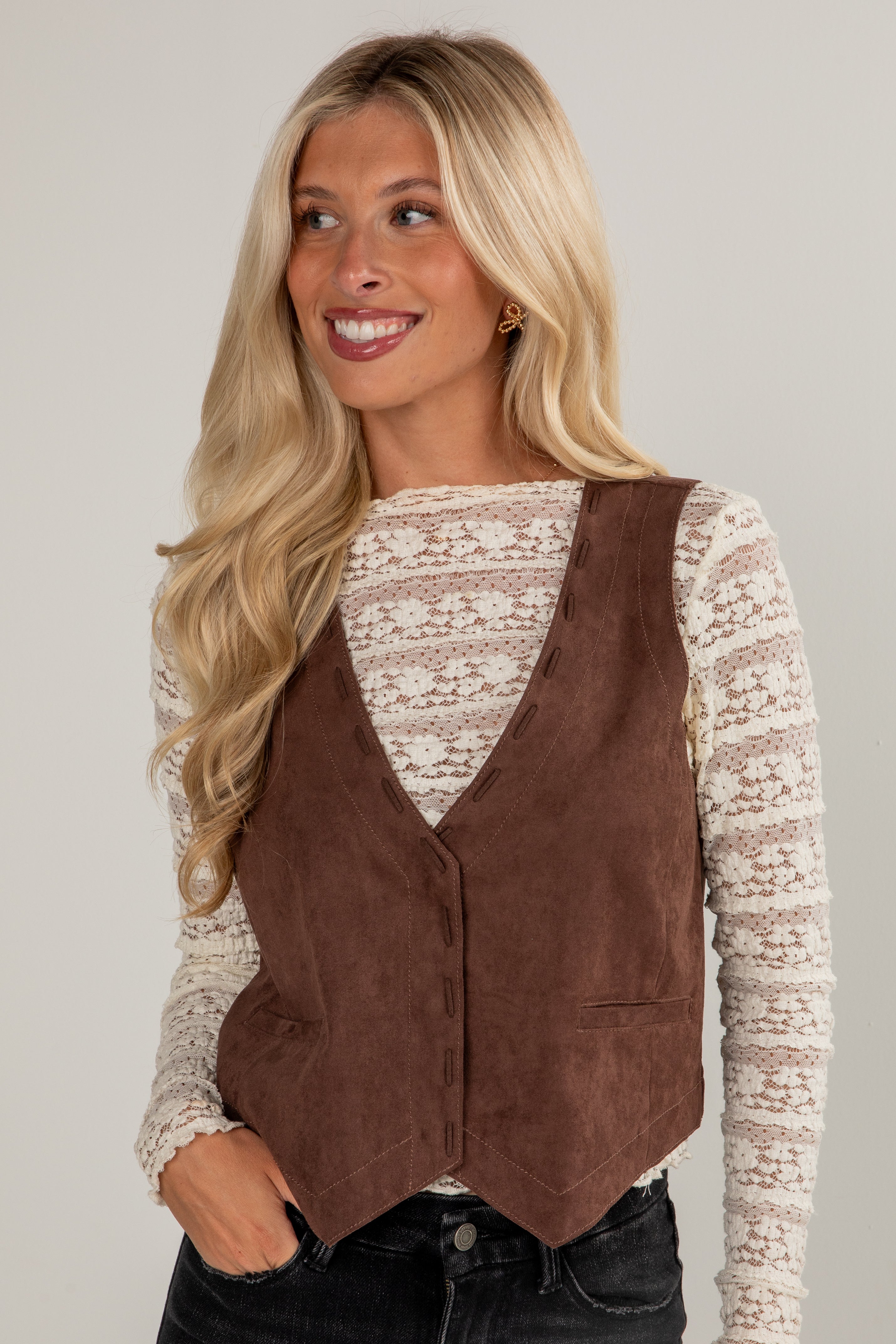Woman wearing a brown vest over a lace top on a plain background
