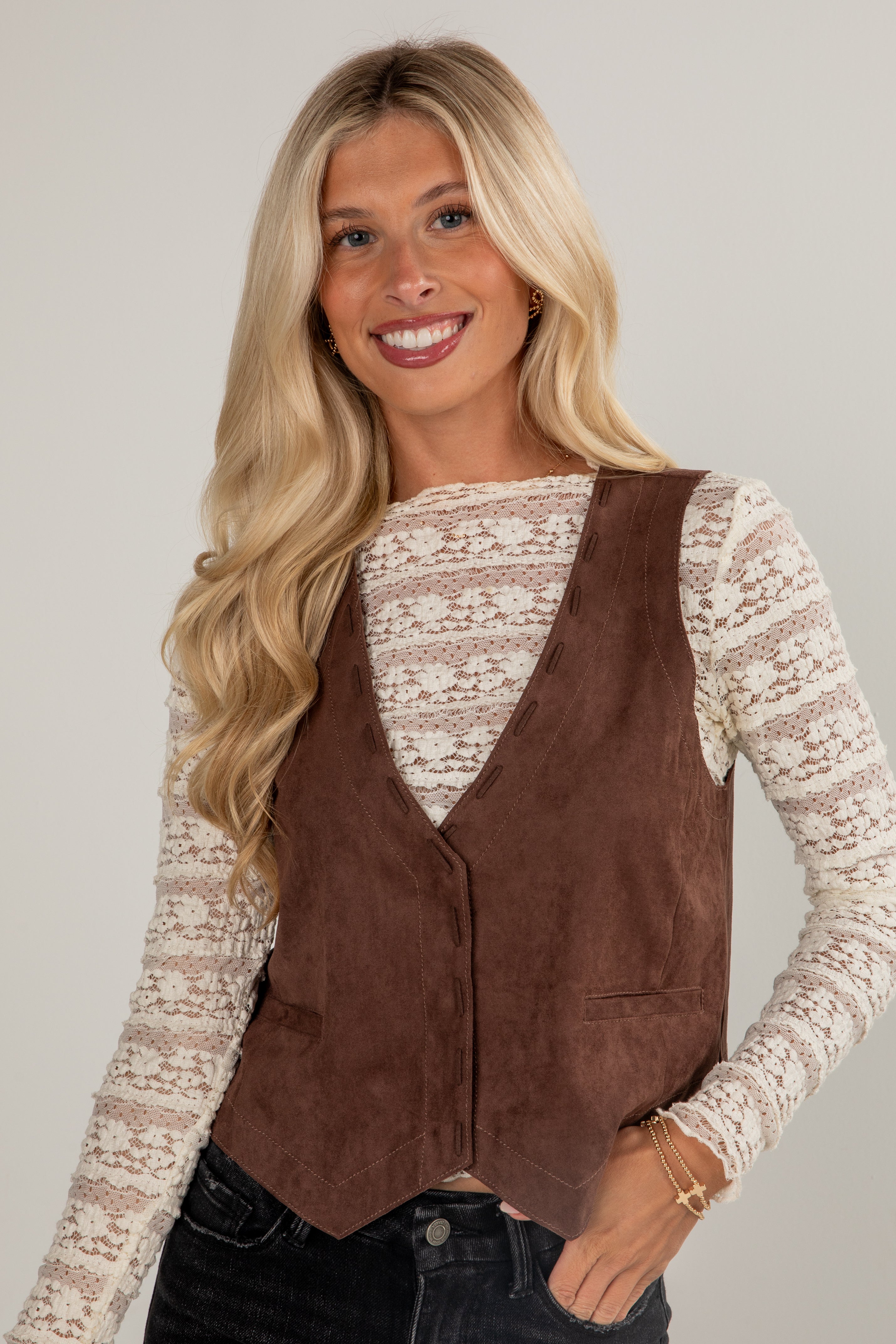 Woman wearing a brown suede vest over a lace top against a plain background