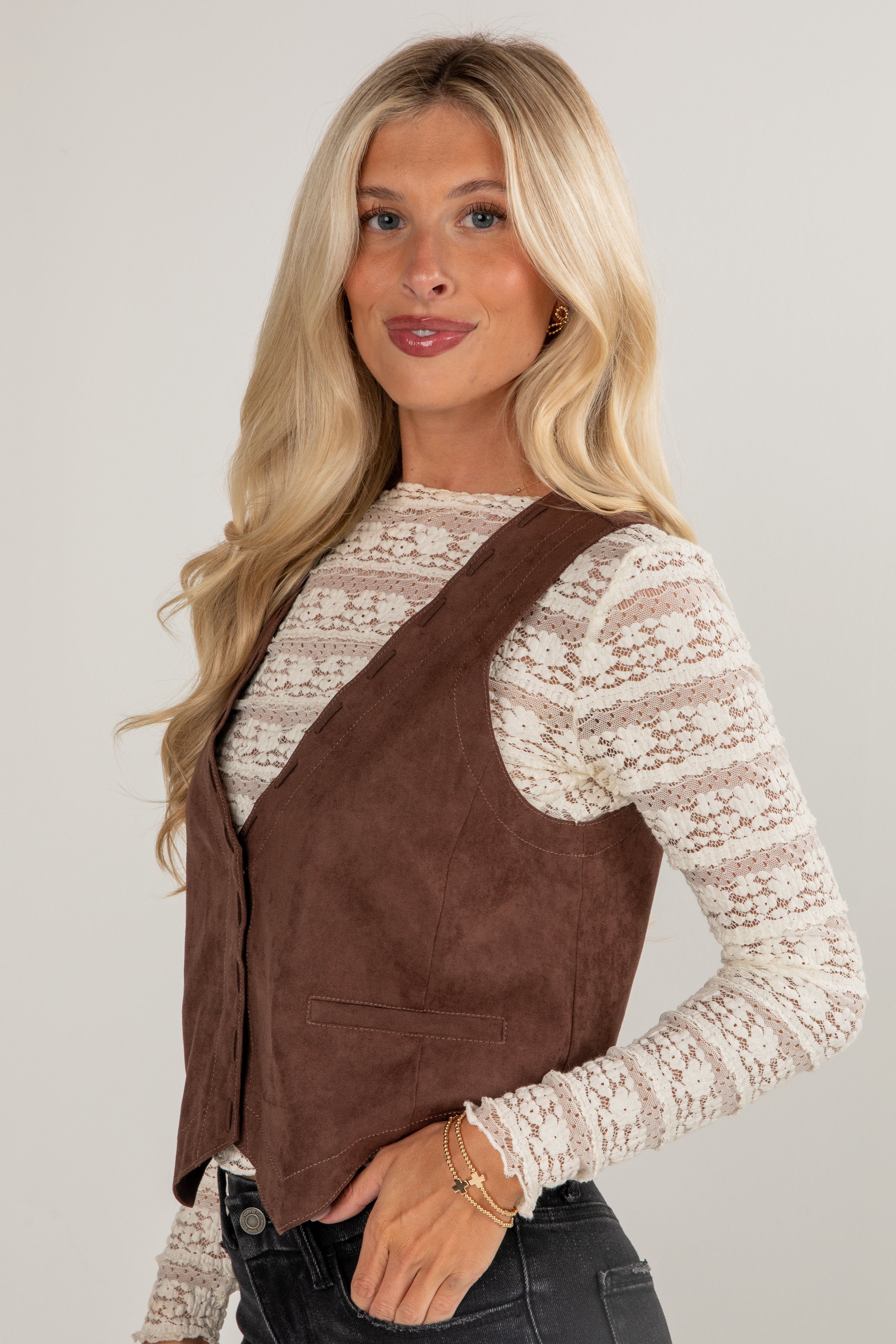 Woman wearing a brown suede vest over a white lace top on a plain background