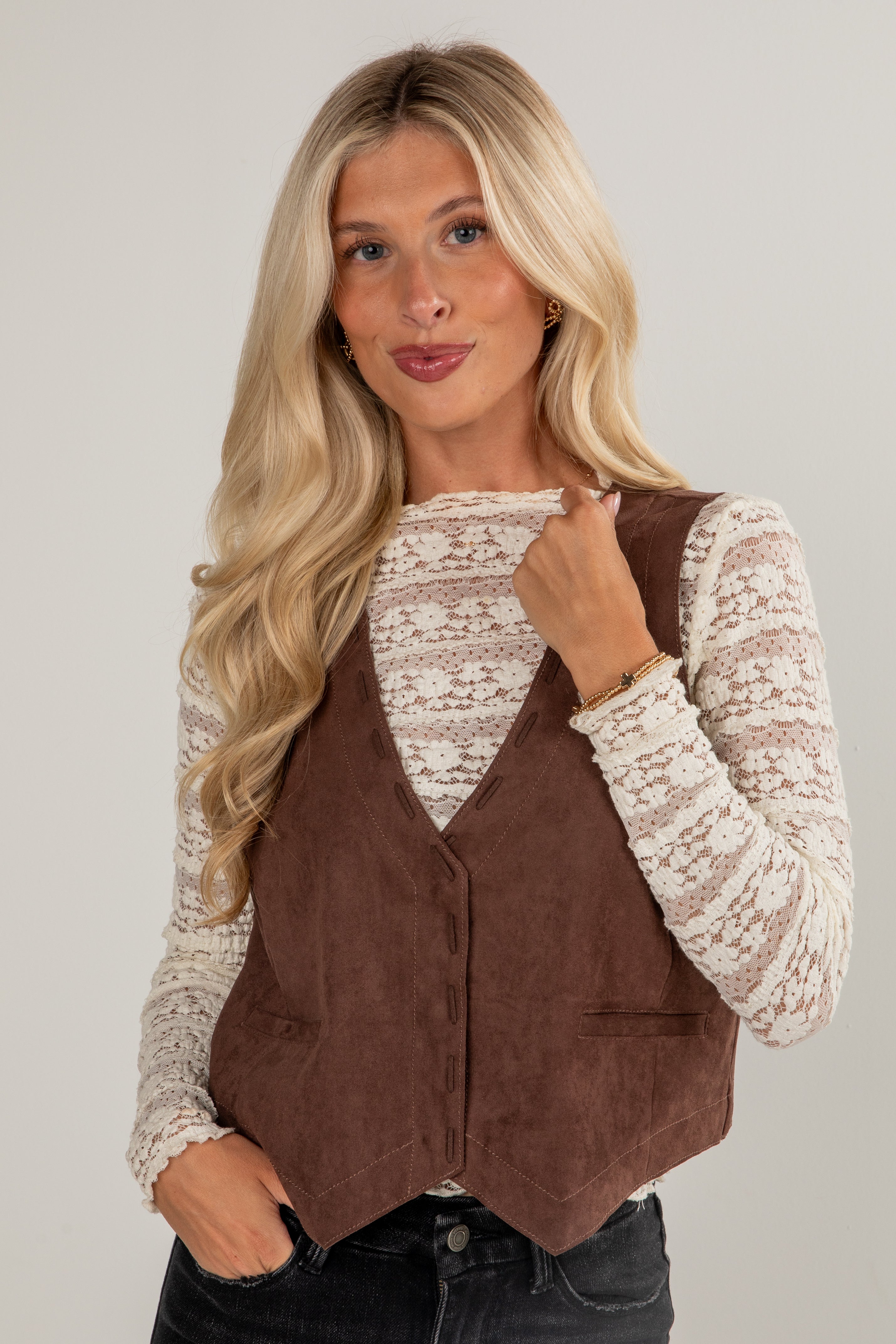 Woman wearing a brown vest over a lace top on a plain background