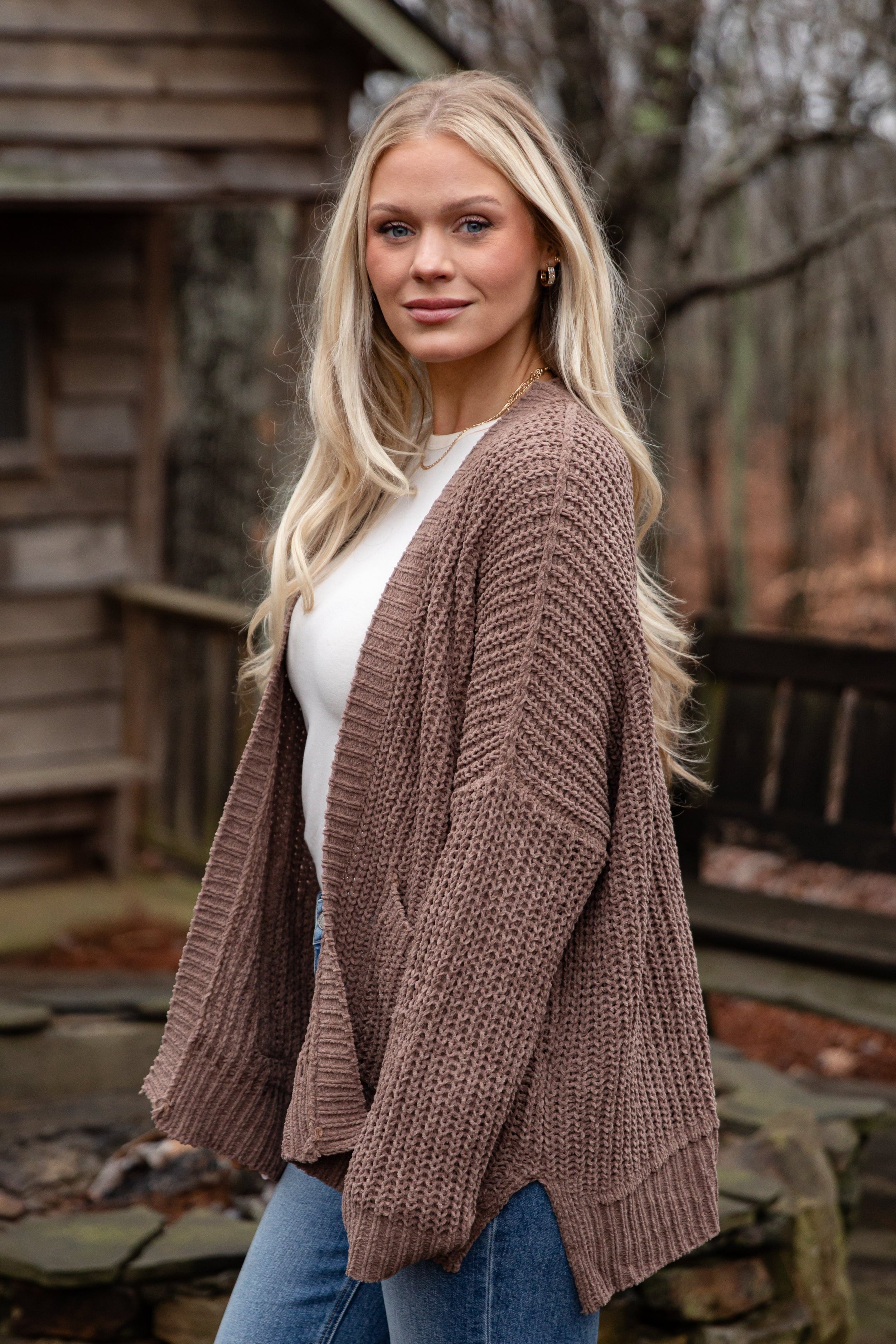 Woman wearing a brown cardigan over a white shirt and blue jeans, standing outdoors.