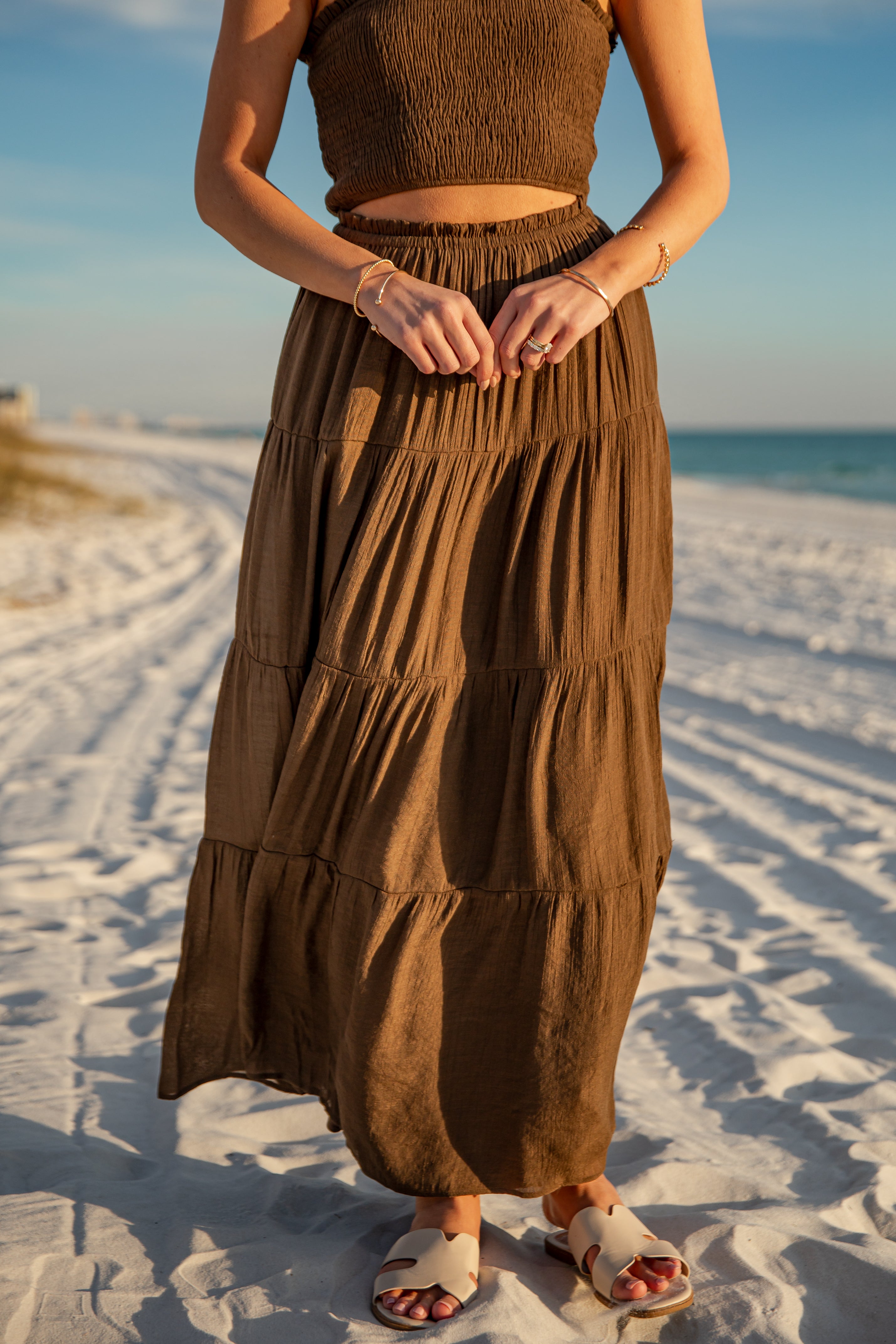 Person wearing a brown dress standing on a sandy beach with ocean in the background