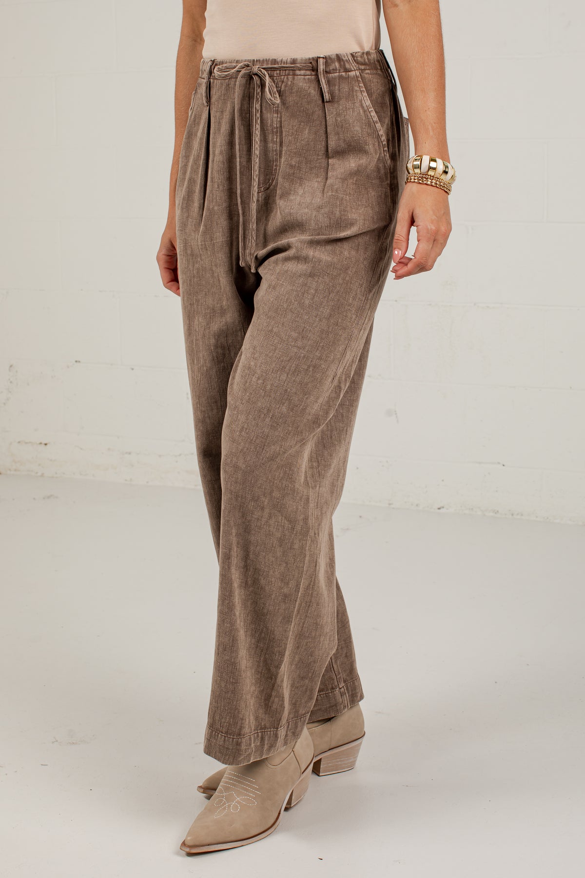 A person wearing high-waisted, wide-leg taupe-brown corduroy pants with a drawstring waist, paired with beige pointed-toe heeled boots against a white studio background.