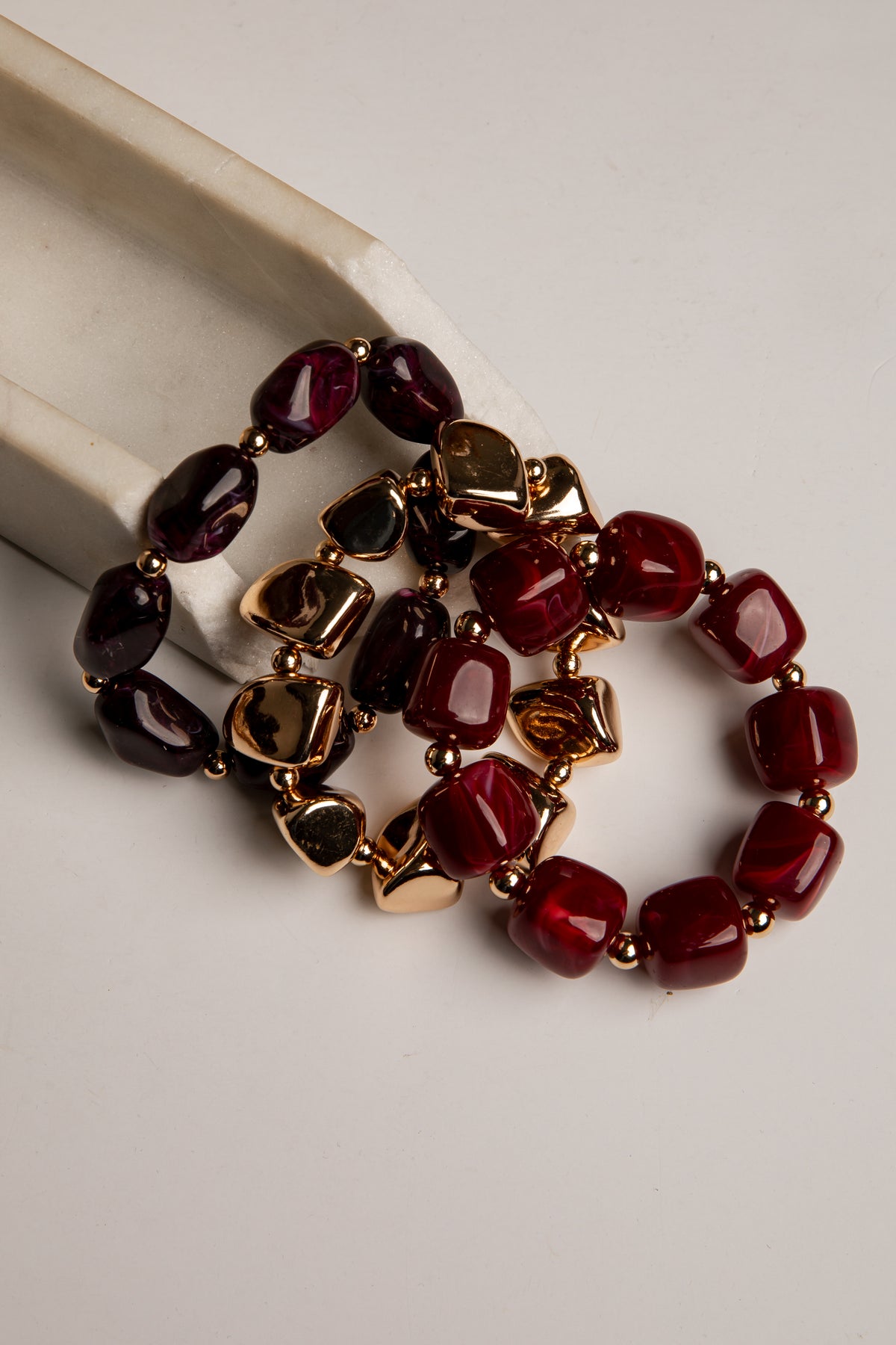 Three beaded stretch bracelets with dark red, burgundy, and black faceted beads separated by gold-toned spacers, displayed on a light beige surface.