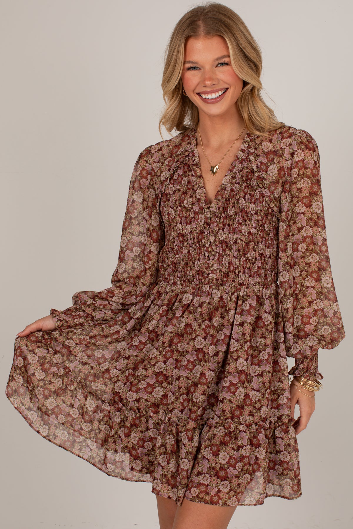 A woman modeling a long-sleeve, smocked-waist mini dress with a brown, pink, and beige floral pattern against a solid light gray background.