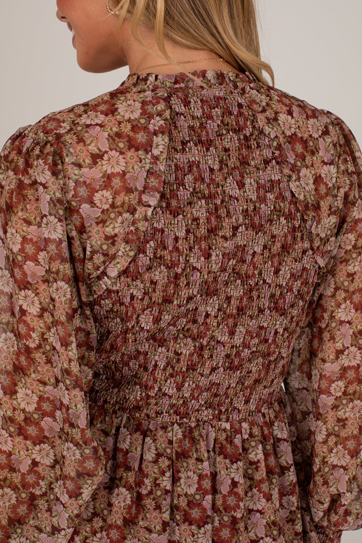 Close-up back view of a woman wearing a long-sleeve smocked dress with a dense floral pattern in dusty pink, rust red, and beige tones against a plain light background.