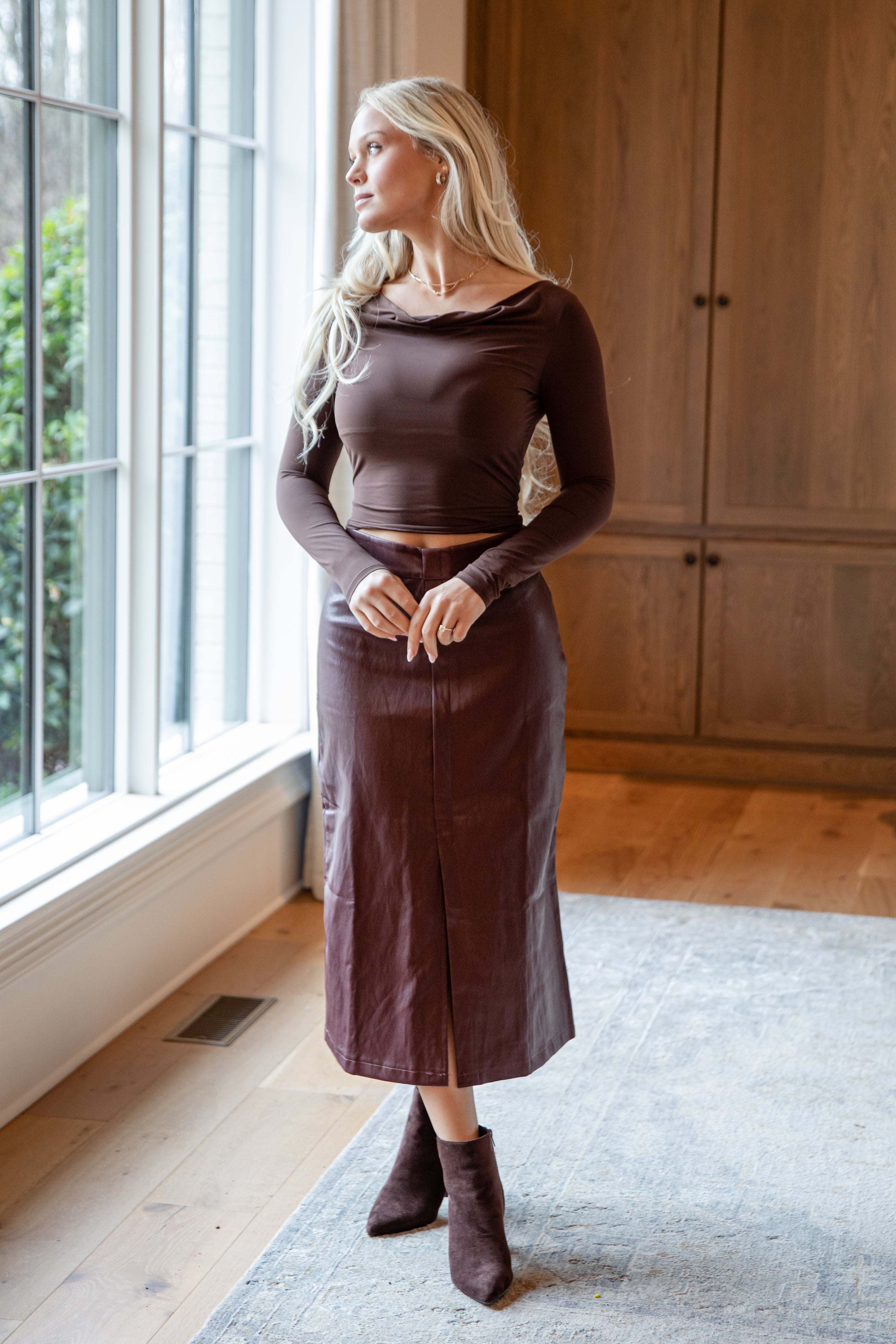 Woman wearing a brown top and skirt standing in a room with wooden flooring and a large window.