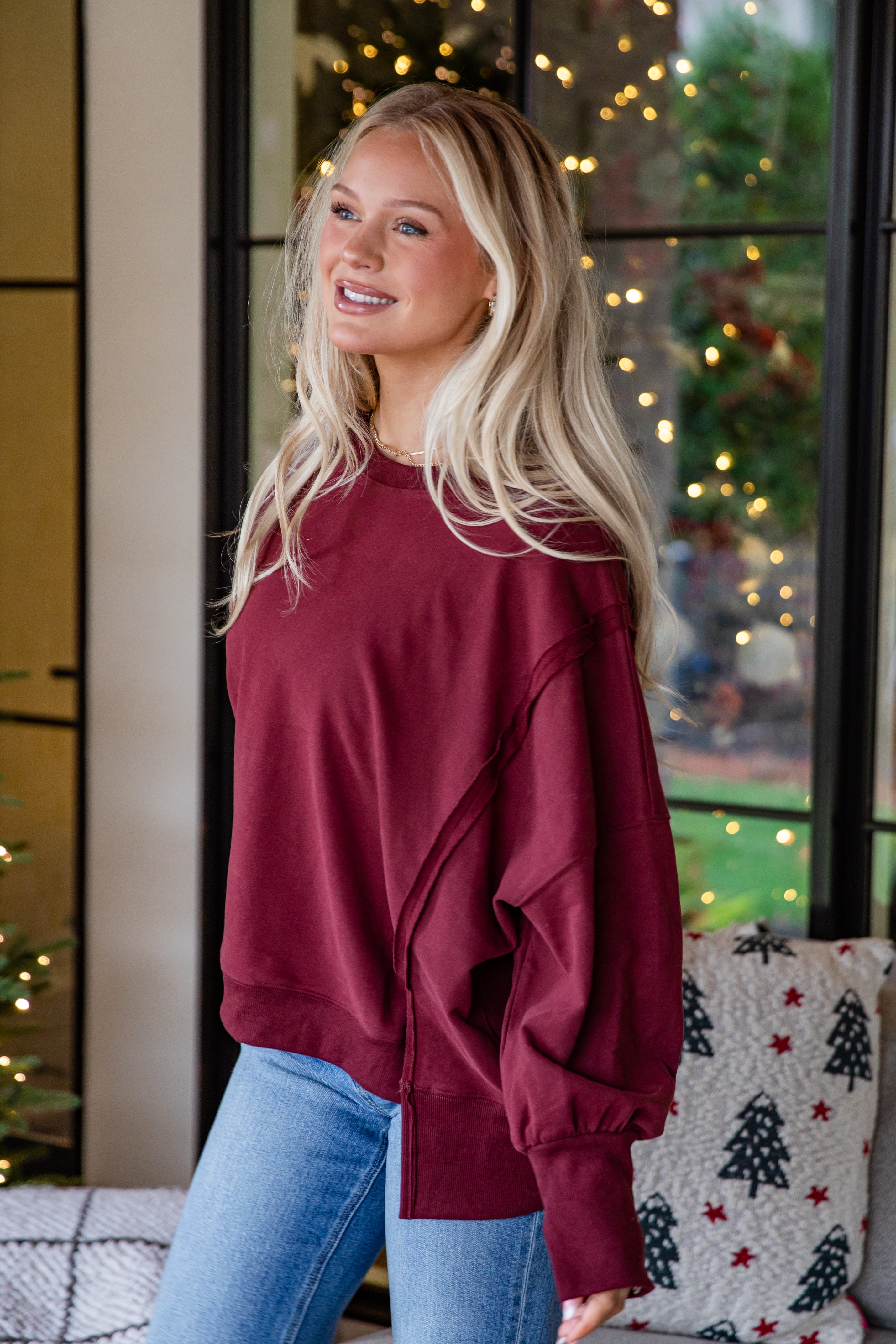 Woman wearing a maroon sweatshirt and blue jeans standing in front of a decorated window with lights.