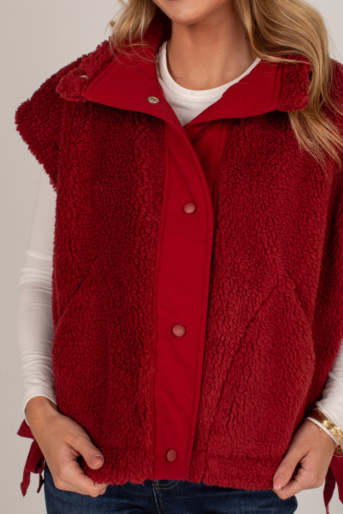 A close-up of a woman wearing a sleeveless red sherpa fleece vest over a white long-sleeve shirt and blue jeans, against a plain light gray background.