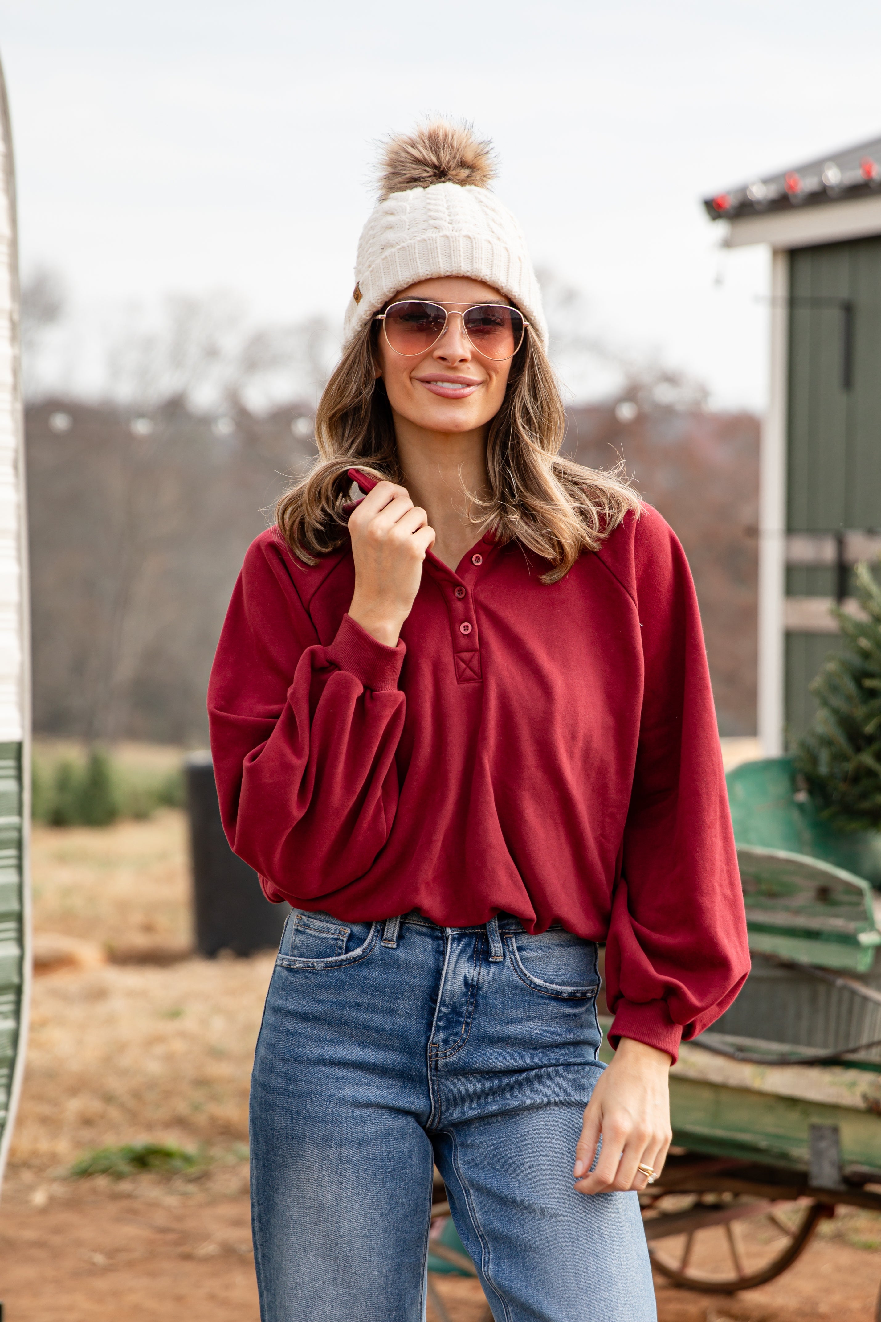 Woman wearing a red shirt, blue jeans, and a white knit hat with a pom-pom outdoors.