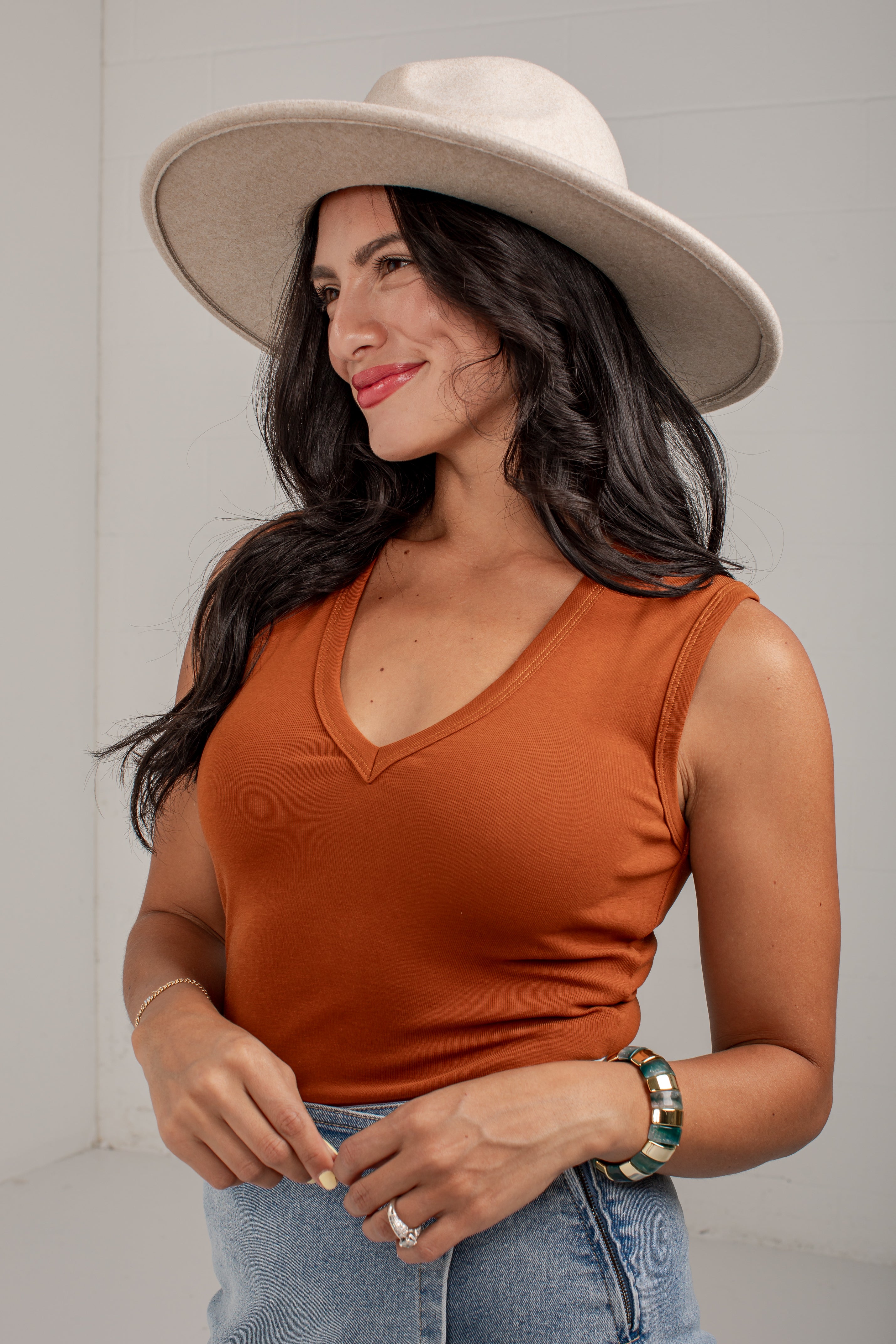 A woman wearing a rust-orange sleeveless V-neck top, a wide-brimmed cream hat, and light-wash blue jeans, standing against a light gray wall.