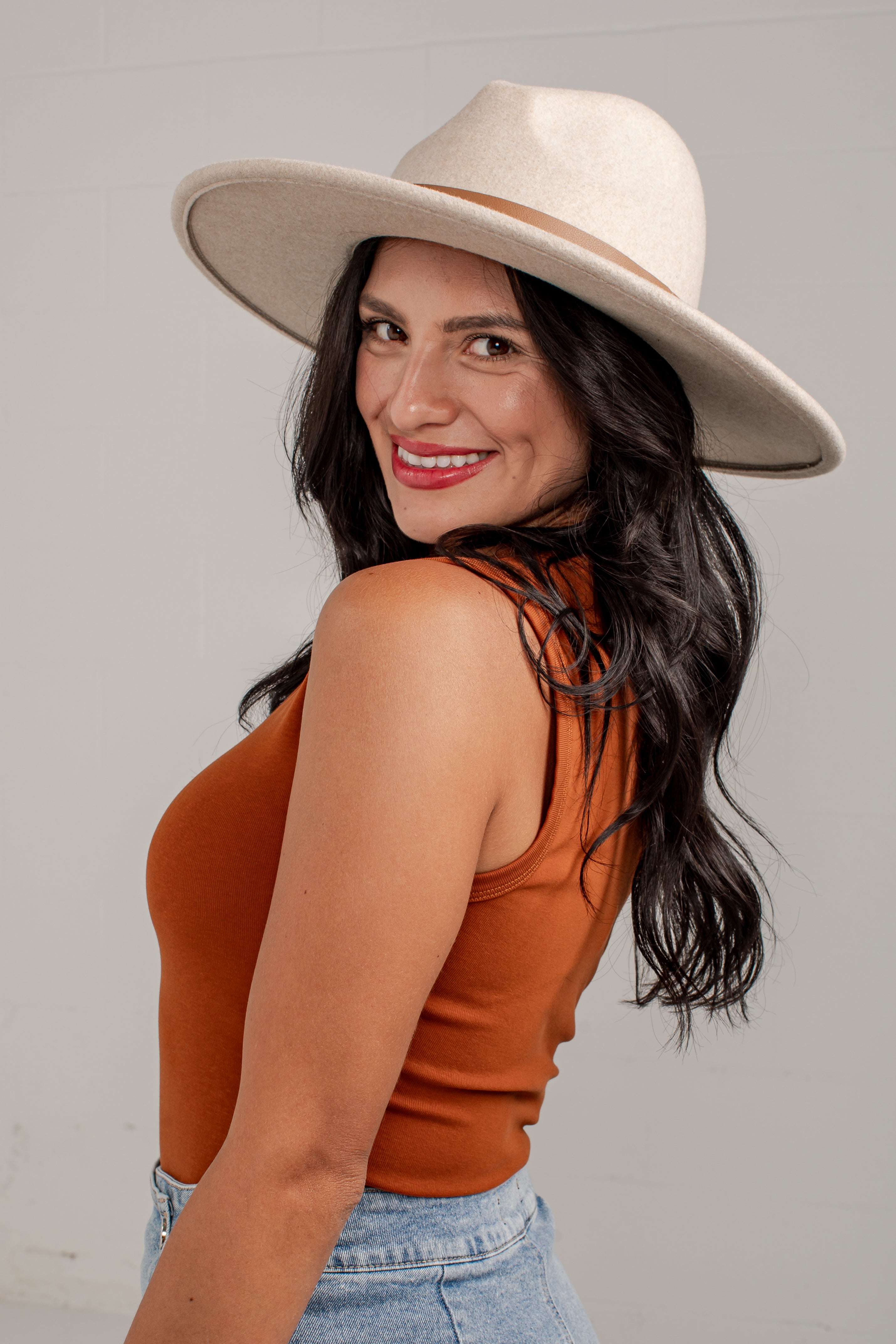 A woman wearing a wide-brimmed cream fedora hat and a rust-orange ribbed tank top, photographed against a light gray studio background.