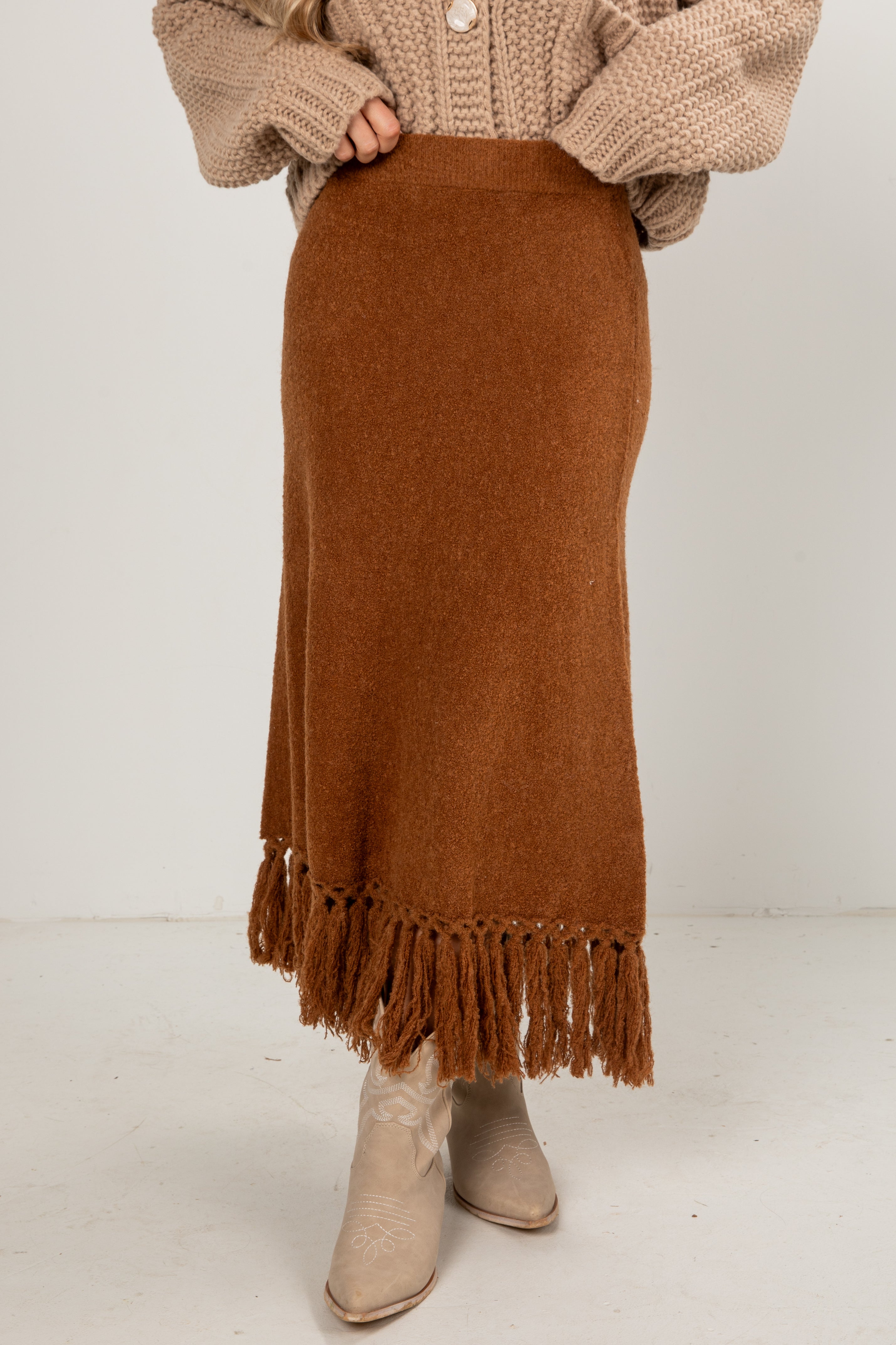 Person wearing a brown fringed skirt with a beige sweater and boots on a white background