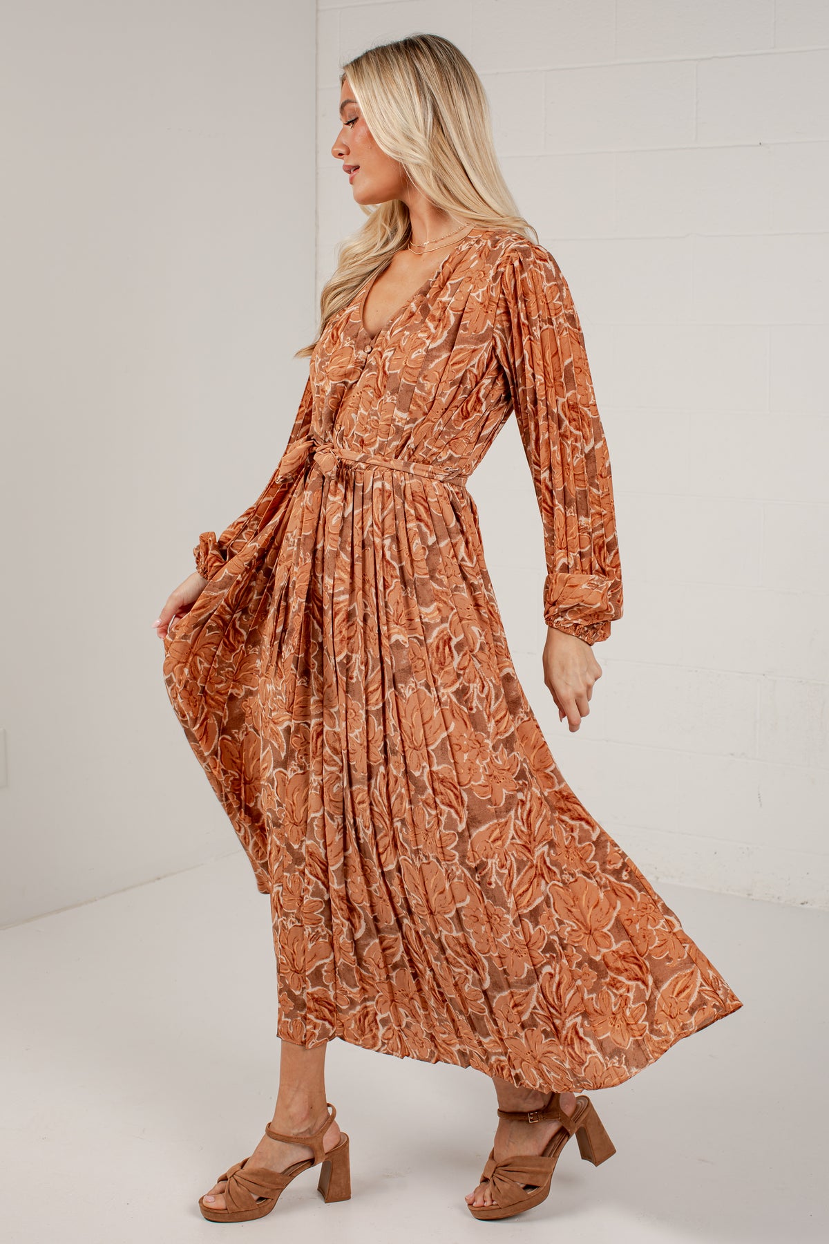 A woman modeling a long-sleeve, wrap-style midi dress with a pleated skirt and an abstract floral pattern in terracotta orange and brown, paired with brown heeled sandals against a white studio background.