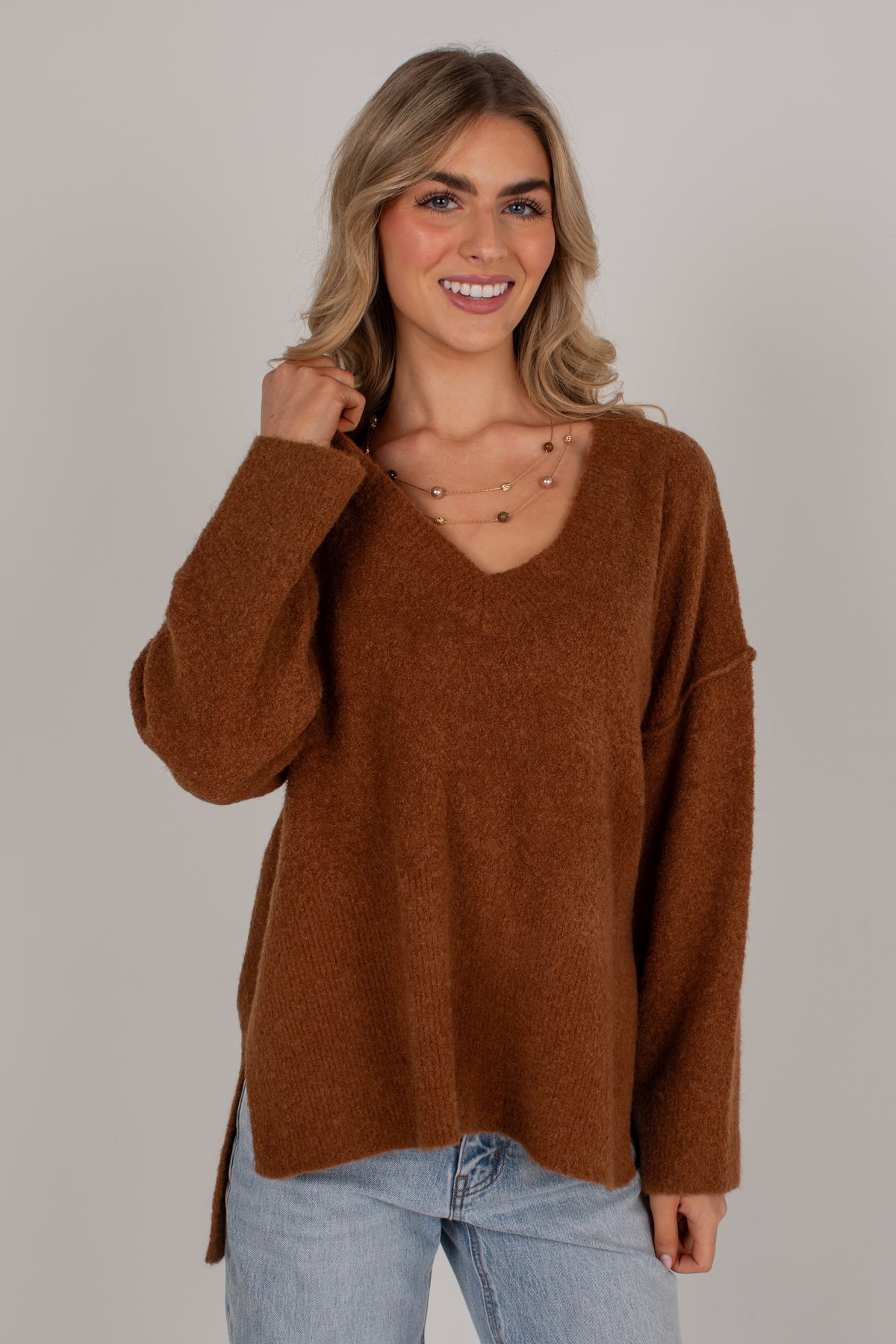 A smiling blonde woman wearing a fuzzy brown V-neck sweater and light blue jeans, standing against a plain light gray studio background.
