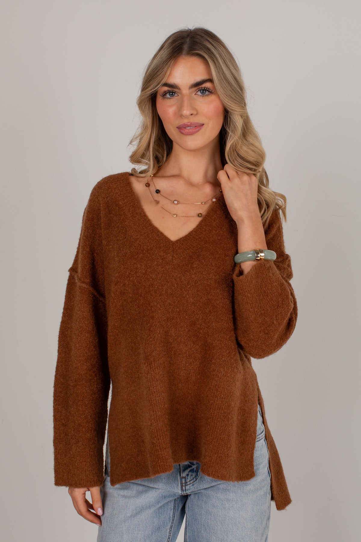 A woman modeling a fuzzy, long-sleeve brown V-neck sweater with side slits, paired with light-wash blue jeans, against a solid light gray background.